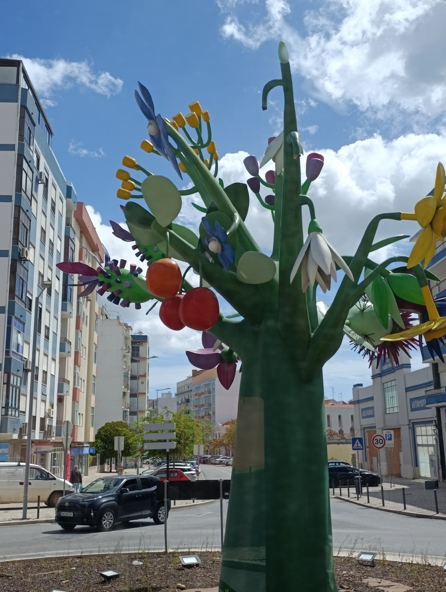 This is a medium-angle, eye-level shot of a colorful, stylized tree sculpture located in Setúbal, Portugal. The sculpture is green with various colorful flower and fruit-like decorations in shades of orange, yellow, blue, and purple. It appears to be made of metal or plastic and is situated on a street corner. In the background, there are modern buildings with white, blue, and pink facades, typical of urban architecture. The street is paved, and there are cars parked and driving. The sky is blue with scattered white clouds, indicating a bright, sunny day.
Specifically, this sculpture is located on Rua 5 de Outubro in Setúbal. This street is a main thoroughfare in the city, characterized by its commercial buildings and pedestrian traffic. The buildings in the background of the image are consistent with the architectural style found in the commercial and residential areas of Rua 5 de Outubro. The presence of street signs, including a "30" speed limit sign and a pedestrian crossing sign, further confirms the urban street environment. A black car with license plate BO 90 ZR is prominently visible in the foreground, driving on the street. The overall scene captures a moment in the daily life of this Portuguese city, blending public art with its urban landscape.