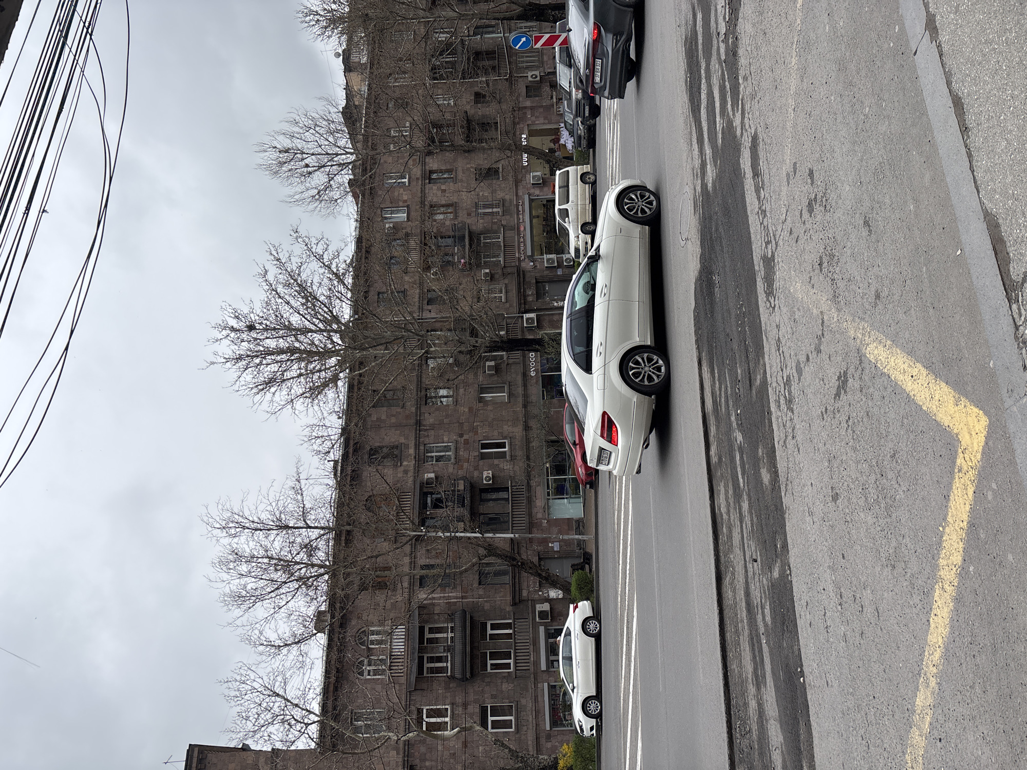 The image captures a street scene in Yerevan, Armenia. The primary subjects are parked cars, including a white sedan and a gray vehicle further down the street. A multi-story building with a weathered stone facade occupies the background, its windows, balconies, and aging air conditioning units adding to the urban texture. Bare trees line the street, their branches contrasting against the overcast sky. Power lines stretch across the upper left corner of the frame. The street is paved and marked with yellow lines. There is no visible human activity in the image.