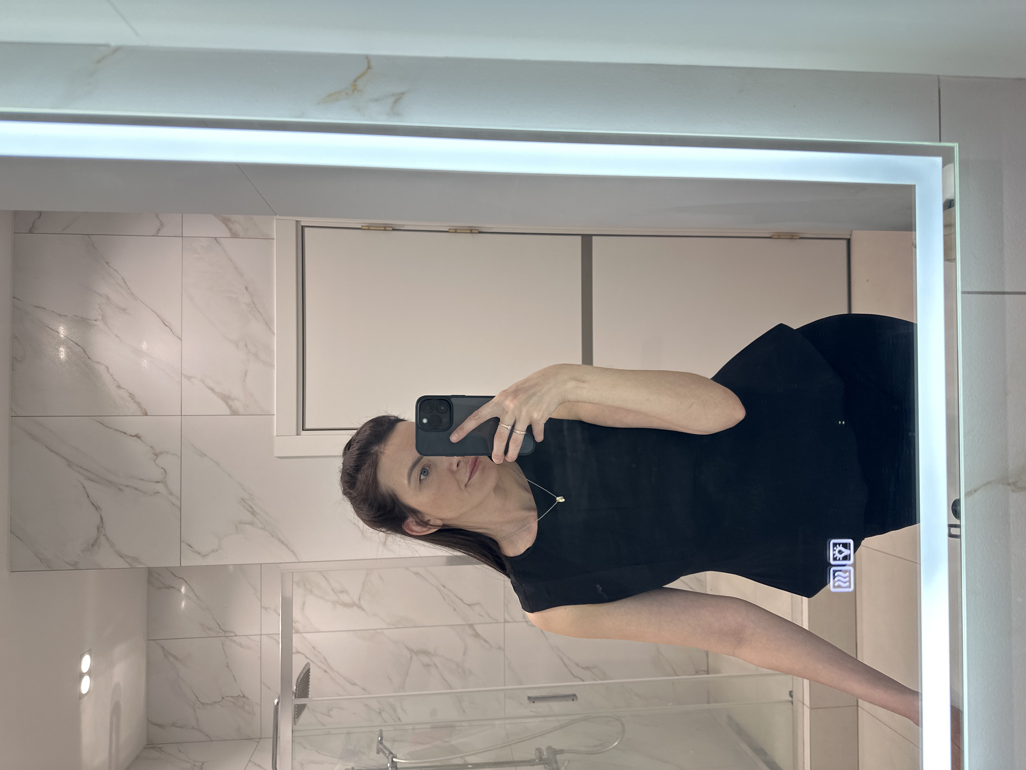 A person is taking a selfie in a bathroom with a modern, illuminated mirror. The bathroom features marble-patterned tiles and a glass-enclosed shower. The person is wearing a black top and a necklace. The location is Прикамский, Russia.