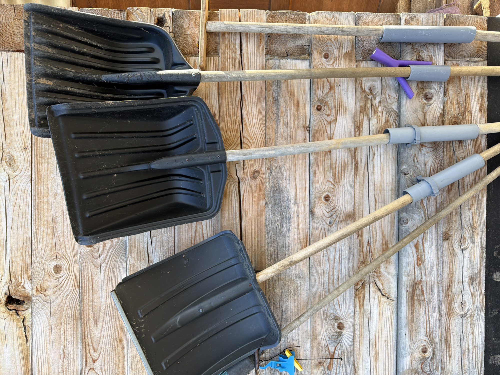 The image shows three black snow shovels leaning against a wooden wall. The shovels have wooden handles with grey plastic grips. One handle has a purple attachment. The wall is made of rough-hewn wooden planks. The scene is likely located in a storage area or garage based on the setting, and is consistent with locations in Фоки, Russia.