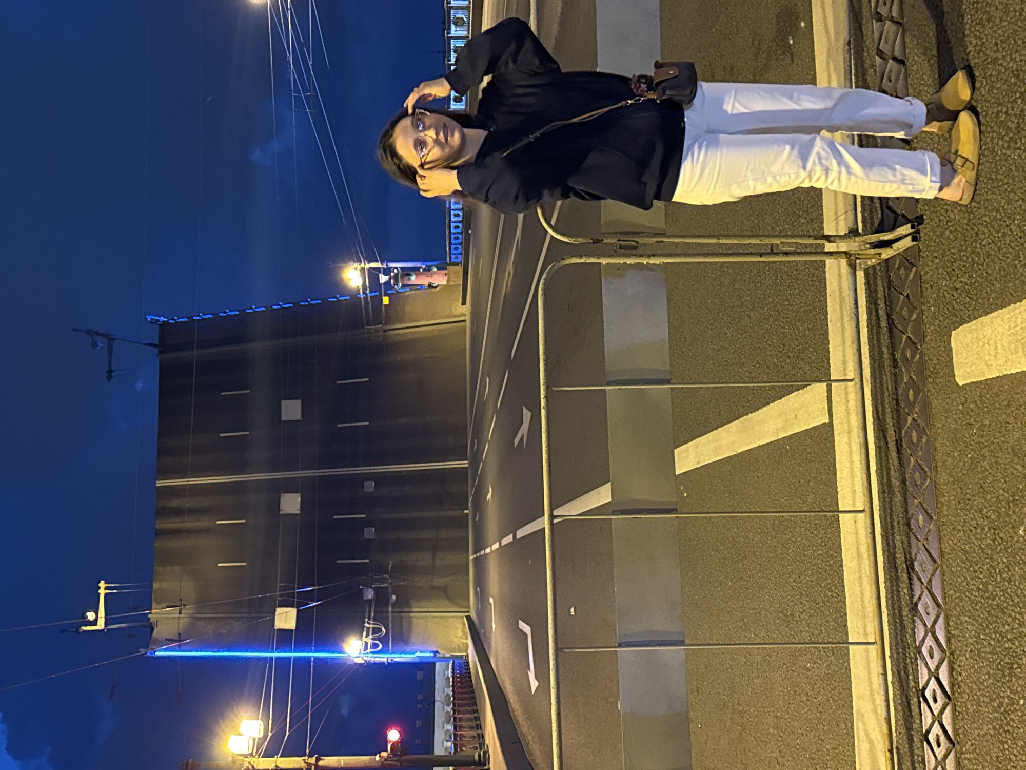 A young woman stands on a bridge in St. Petersburg, Russia at night. The bridge is raised, revealing a dark, upward-sloping road. The woman is wearing glasses, a navy blue hoodie, white pants, and yellow slip-on shoes. She has a brown crossbody bag. She is looking at the camera and touching her hair. The bridge has traffic lights, streetlights, and blue neon lights. The sky is dark blue with some clouds. The road has white lane markings and arrows. There are metal barriers in the foreground.