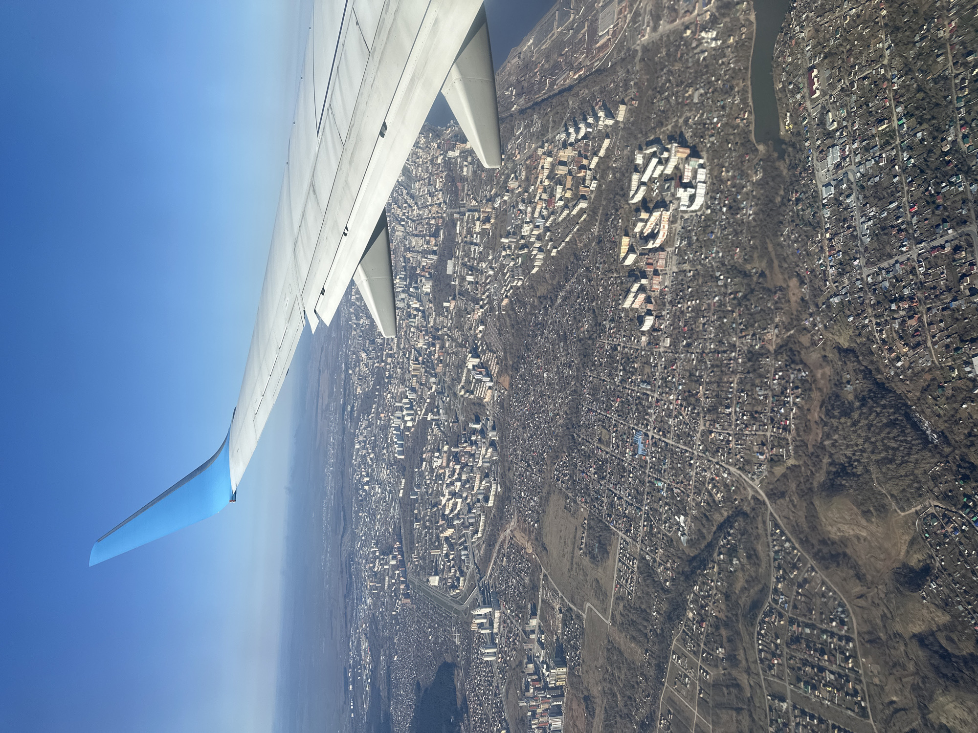 An aerial view from an airplane window shows the city of Sokol, Russia. The wing of the plane is visible in the foreground, with the city spread out below. The urban landscape features a mix of residential buildings, roads, and patches of trees. A river runs through the city. The sky is a clear blue.
