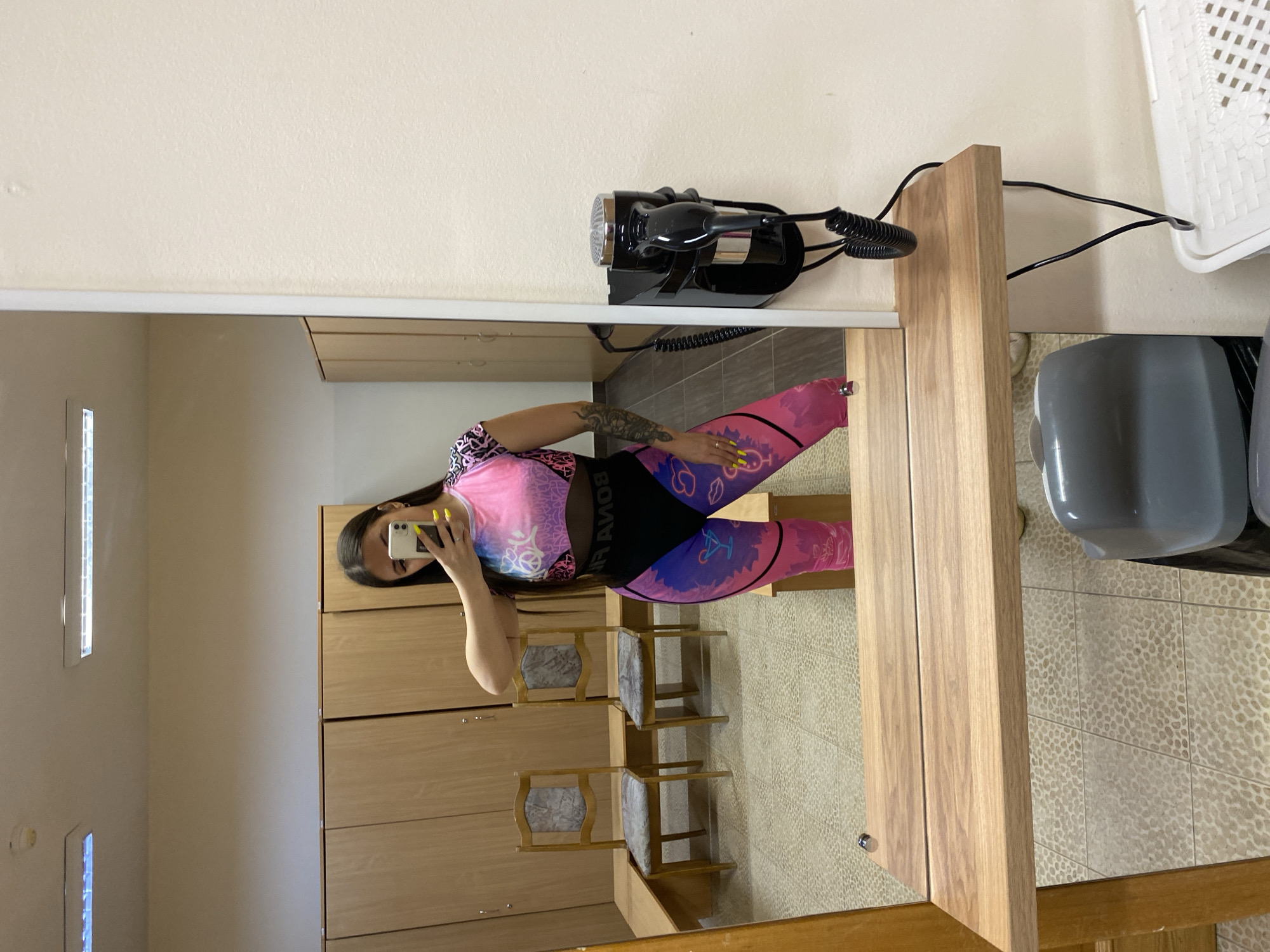The image appears to be a mirror selfie taken in a room with a dressing area or bathroom. The reflection shows a person holding a smartphone, likely taking the picture. The individual is wearing a colorful patterned athletic outfit consisting of a top and leggings. Behind the person are wooden cabinets or lockers, and there are small chairs with cushioned seats. There is a hairdryer mounted on the wall, along with other bathroom fixtures. The floor is tiled. The location given is Chaikovsky, Russia. There is no readily identifiable location in the city that would further describe the image.