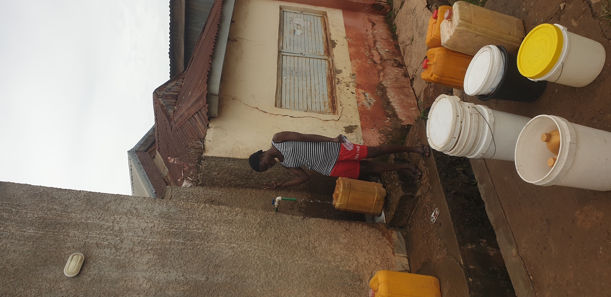 In Sabon Tasha, Nigeria, an African female stands beside a rough-textured, weathered concrete wall, a blue-handled faucet attached to a green pipe on the wall. Water streams from the faucet into a large, opaque yellow plastic container positioned on a raised step. The woman, wearing a striped tank top and red shorts, holds a white plastic bag and makes a peace sign with her left hand. Behind her, a building with a corroded, rusty corrugated metal roof is visible, featuring a cream-colored wall with orange-brown staining and a boarded-up window. In the foreground, several white plastic buckets are stacked, and more yellow and beige plastic containers are arranged on the ground, suggesting water collection. The ground is damp and appears to be a dirt pathway. The sky is overcast.