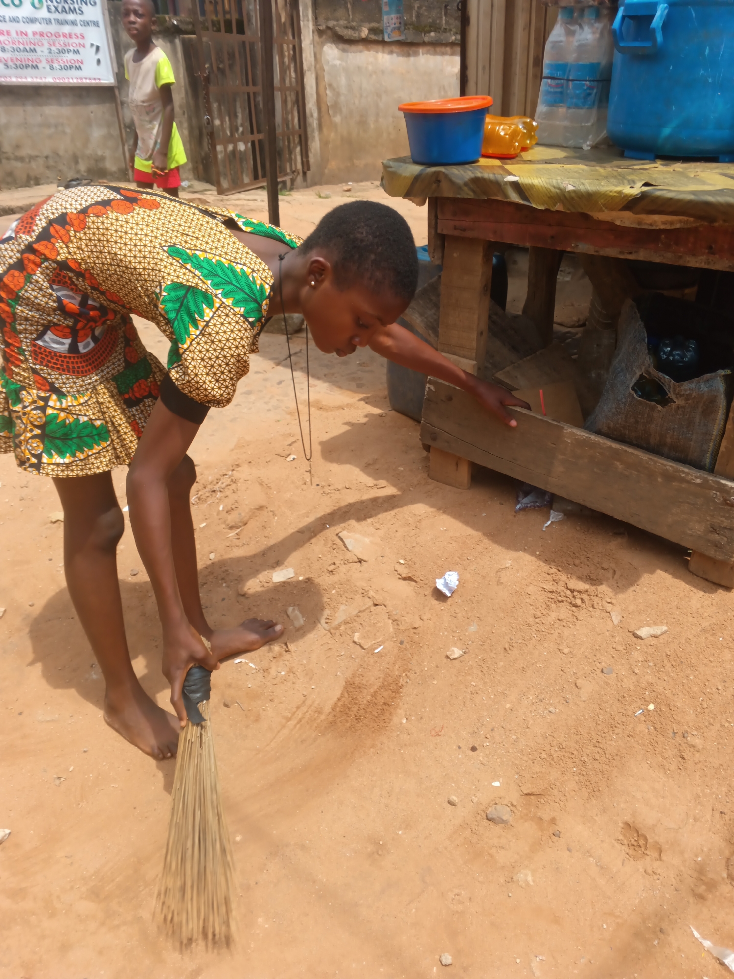 In Onitsha, Nigeria, a young girl with dark skin and short, dark hair is sweeping a dusty, sandy ground with a traditional broom. She is wearing a vibrantly patterned dress with green, orange, and yellow motifs, and a black band on her upper arm. Her body is bent at the waist as she maneuvers the broom. In the background, another child, also with dark skin and short hair, stands near a metal gate. Further back, a building with a faded sign and a weathered texture is visible. To the right, a wooden table is set up with various items, including blue and orange plastic containers, plastic bottles, and a large blue drum. A woven sack is also present under the table. The overall environment appears to be an outdoor marketplace or street scene.