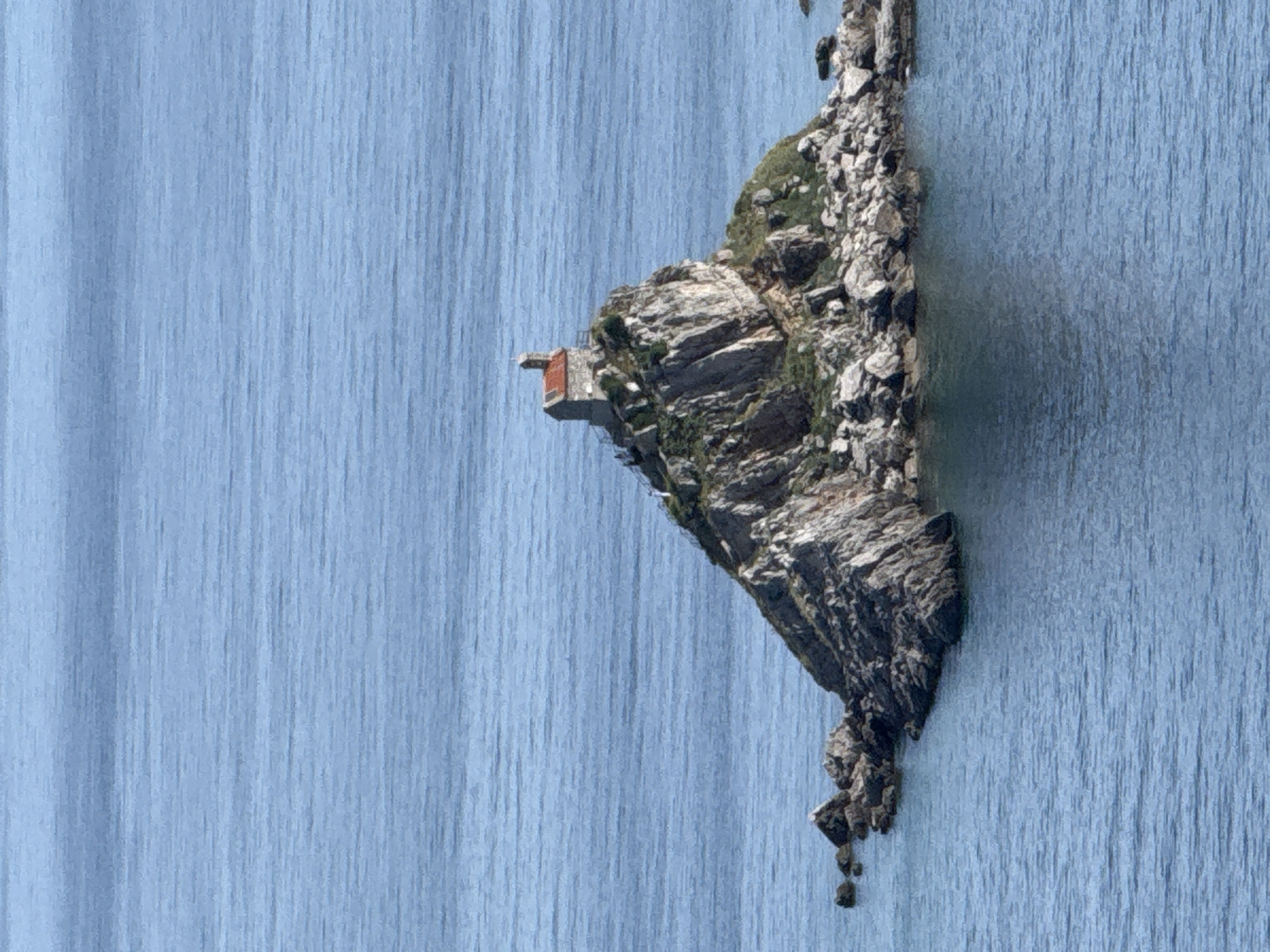 The image shows a rocky islet in Mirište, Montenegro, with a small, modern structure perched on the cliff. The surrounding water appears to be a light blue hue. The islet's rocky terrain features varying textures, with patches of green vegetation. The water's surface exhibits a vertical texture.
