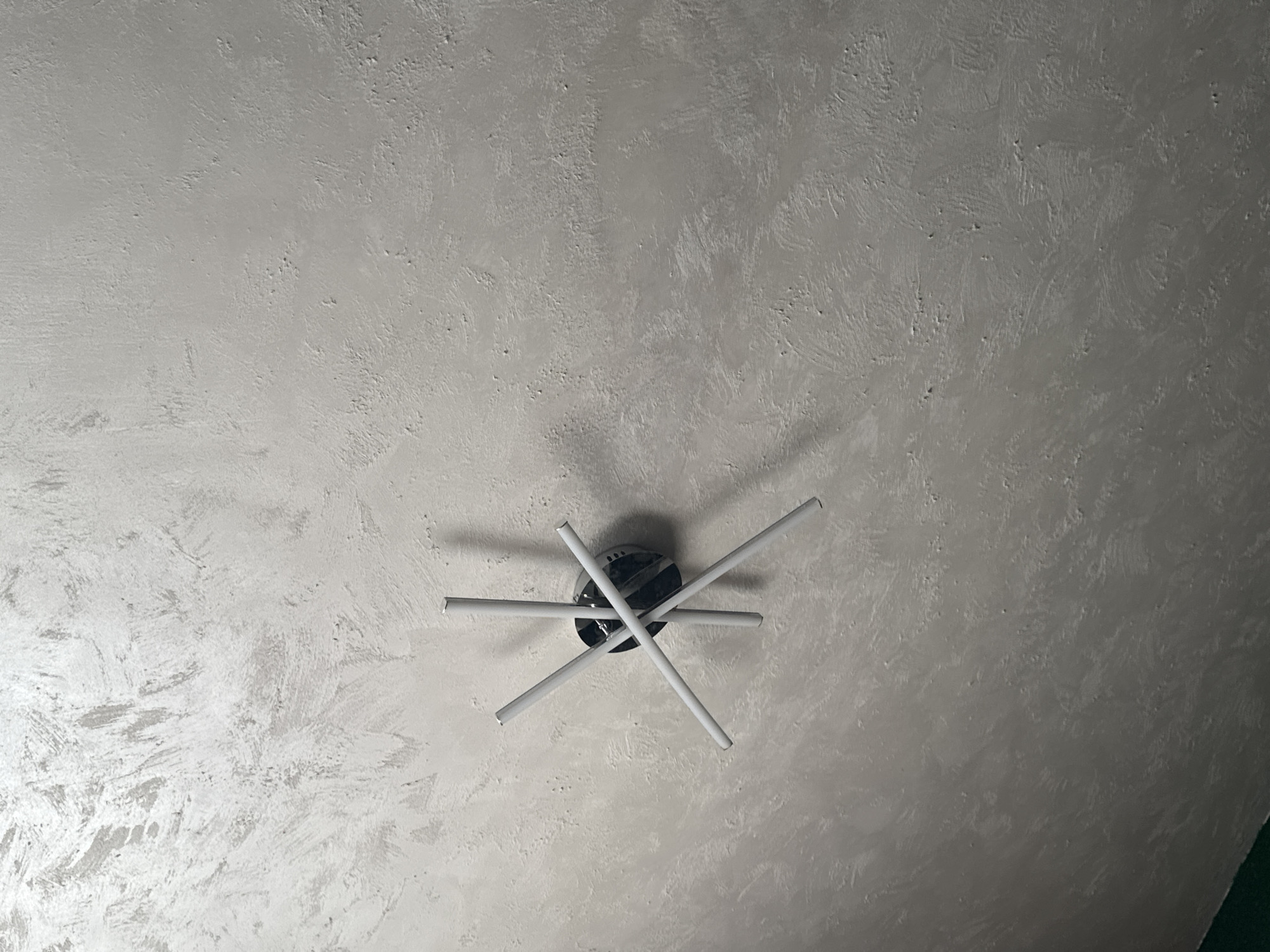 A low-angle shot shows a modern ceiling light fixture against a textured, light gray ceiling. The light fixture has a circular, black base from which four white cylindrical arms extend outwards in a pinwheel arrangement. The ceiling has a mottled appearance with variations in the gray color, suggesting a textured paint or plaster finish. The lighting in the scene is even, and the texture of the ceiling gives it a slightly rough appearance. No people or other objects are visible.