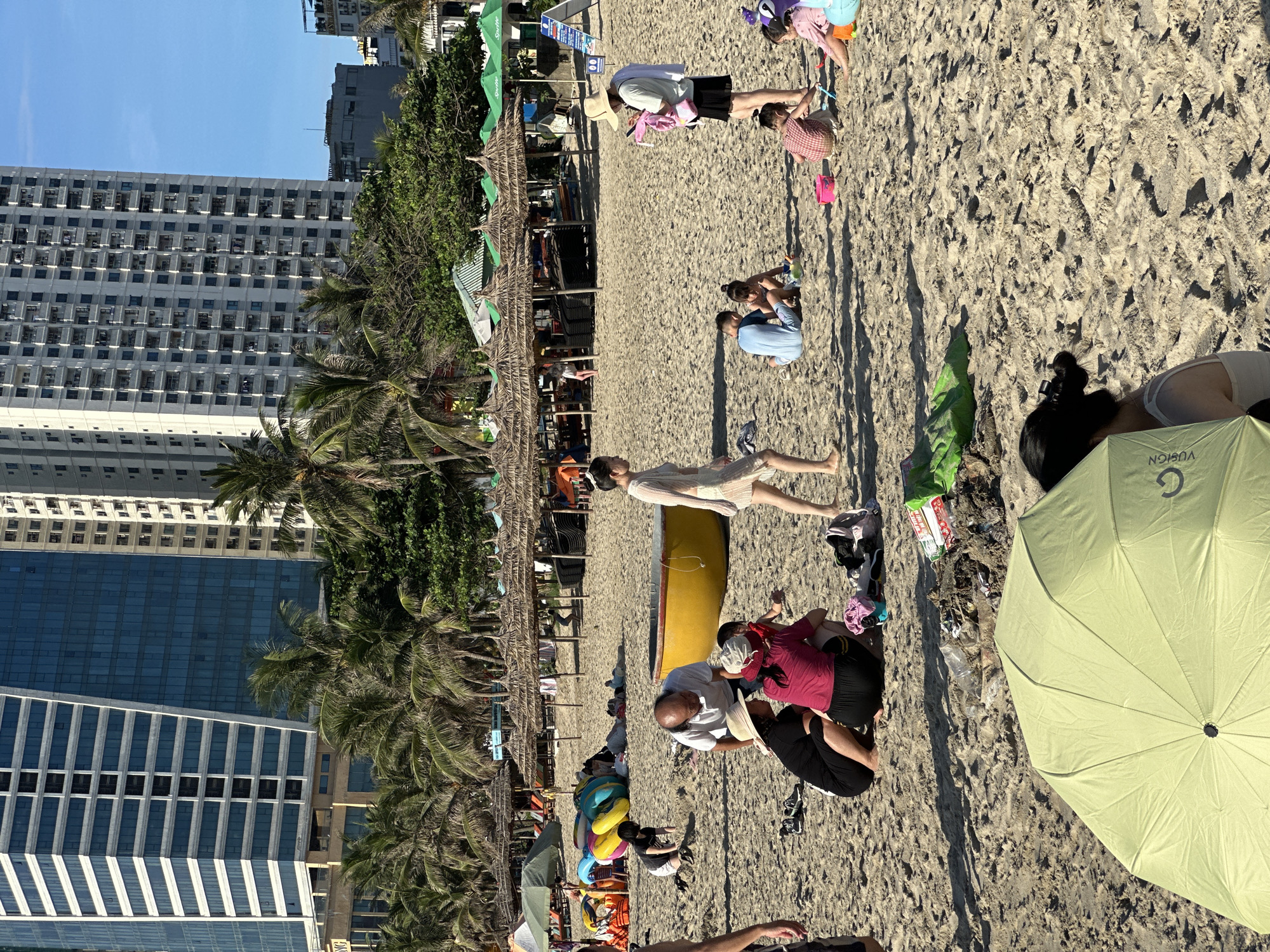 In Đà Nẵng, Vietnam, a sunlit beach scene unfolds with a backdrop of modern high-rise buildings and lush palm trees. The sandy shore is dotted with numerous beachgoers, engaged in various activities. In the foreground, a large, light green umbrella partially obscures a woman. Further back, a group of people are gathered, some sitting on the sand, including a man in a white shirt and several women. A bright yellow, boat-shaped object is visible near them.
Other individuals and families are scattered across the beach. Some are playing with colorful inflatable rings, while children are seen playing in the sand with toys. Several thatched-roof umbrellas provide shade in the middle ground, with lounge chairs and other beach equipment stored beneath them. The sea is not visible in this frame. The overall atmosphere appears relaxed and recreational, characteristic of a popular beach destination.