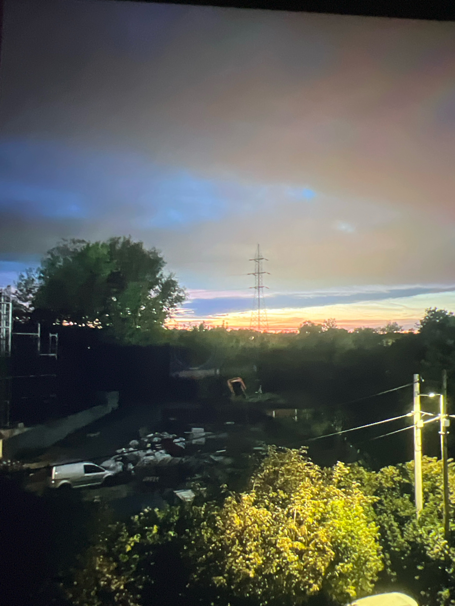 The image captures a twilight scene in the City of Zagreb, Croatia, featuring a landscape dominated by a large, leafy tree on the left and a power transmission tower in the distance. The sky displays a gradient of colors, transitioning from dark gray at the top to blues, oranges, and yellows near the horizon, indicating either sunrise or sunset. In the foreground and mid-ground, there are signs of construction or a work site, including a white van, piles of debris, and what appears to be construction equipment. Trees and other vegetation are visible throughout the scene, with a brightly lit, dense bush or tree on the right side of the image. A utility pole with wires is also present on the right. The overall impression is one of an urban or semi-urban area undergoing development or activity during the transitional light of day.