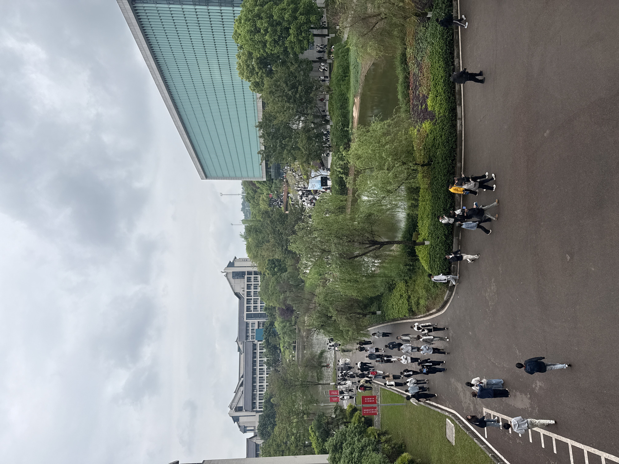 The image presents an aerial view in Jiangjin, China. A body of water divides a walkway and adjacent structures. A large group of people, many wearing black and white attire, is walking along the walkway. There is a large, modern building to the upper left with a grid-like exterior. Along the water, there are trees and dense vegetation. There are multiple buildings across the water, appearing to be part of a complex. Signs with Chinese writing are visible on the lower left. The sky is overcast.