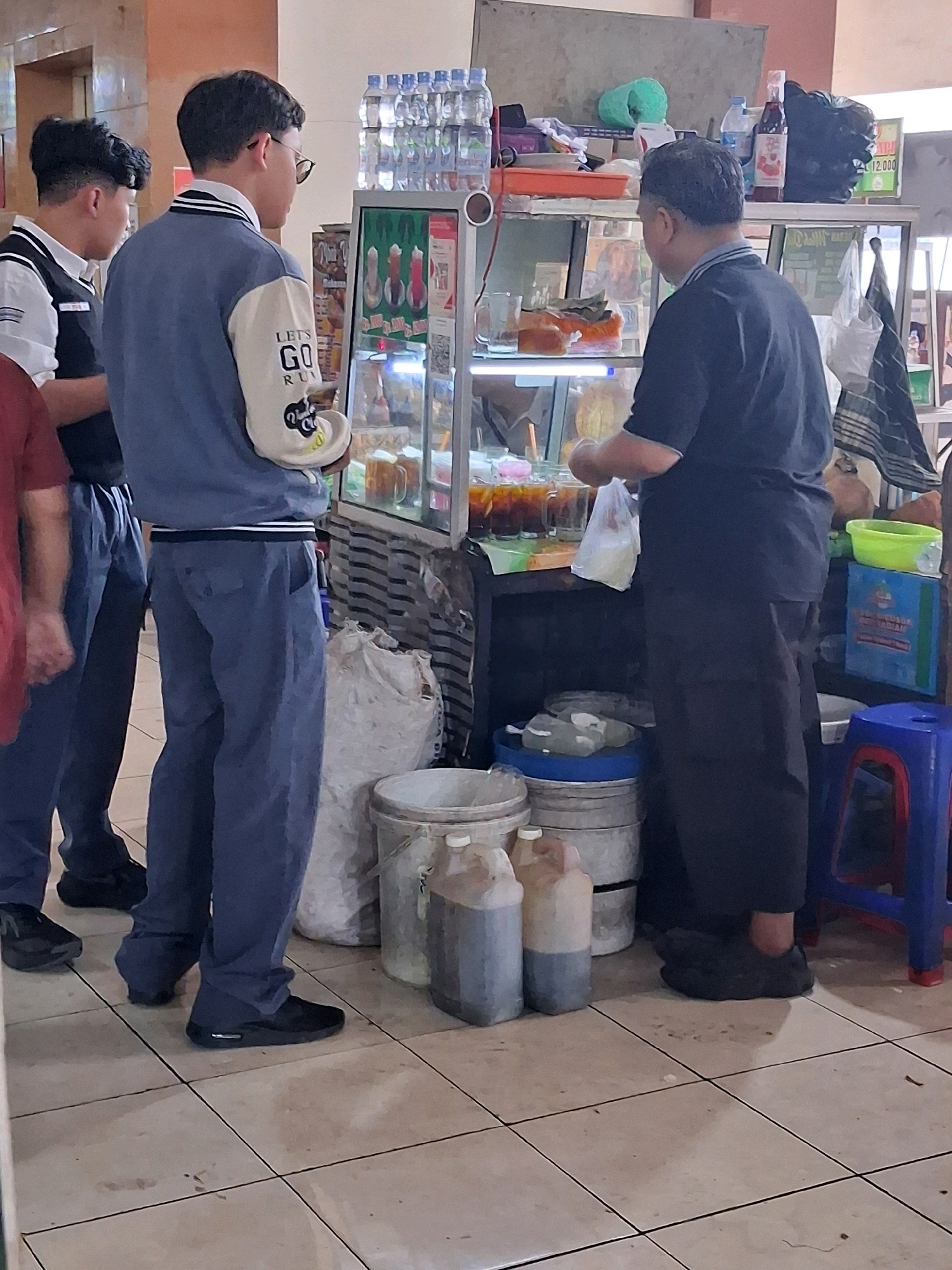 The image captures an outdoor scene in Surakarta, Indonesia, likely a food stall or small eatery. In the foreground, three individuals are present. Two younger males, seemingly students given their attire, stand to the left, facing a display case. One is wearing a blue jacket with "LETS GO RUN" printed on the sleeve. To the right, an adult male, dressed in a dark t-shirt and pants, is actively serving or preparing food, holding a plastic bag.
The environment includes a glass display case showcasing beverages in glasses and bottles. Various containers, including large white buckets and smaller plastic jugs, are arranged on the floor. A blue plastic stool is visible to the right, and a green plastic container sits on a shelf above the display case. The ground is tiled. The overall impression is a bustling, everyday scene at a local vendor.