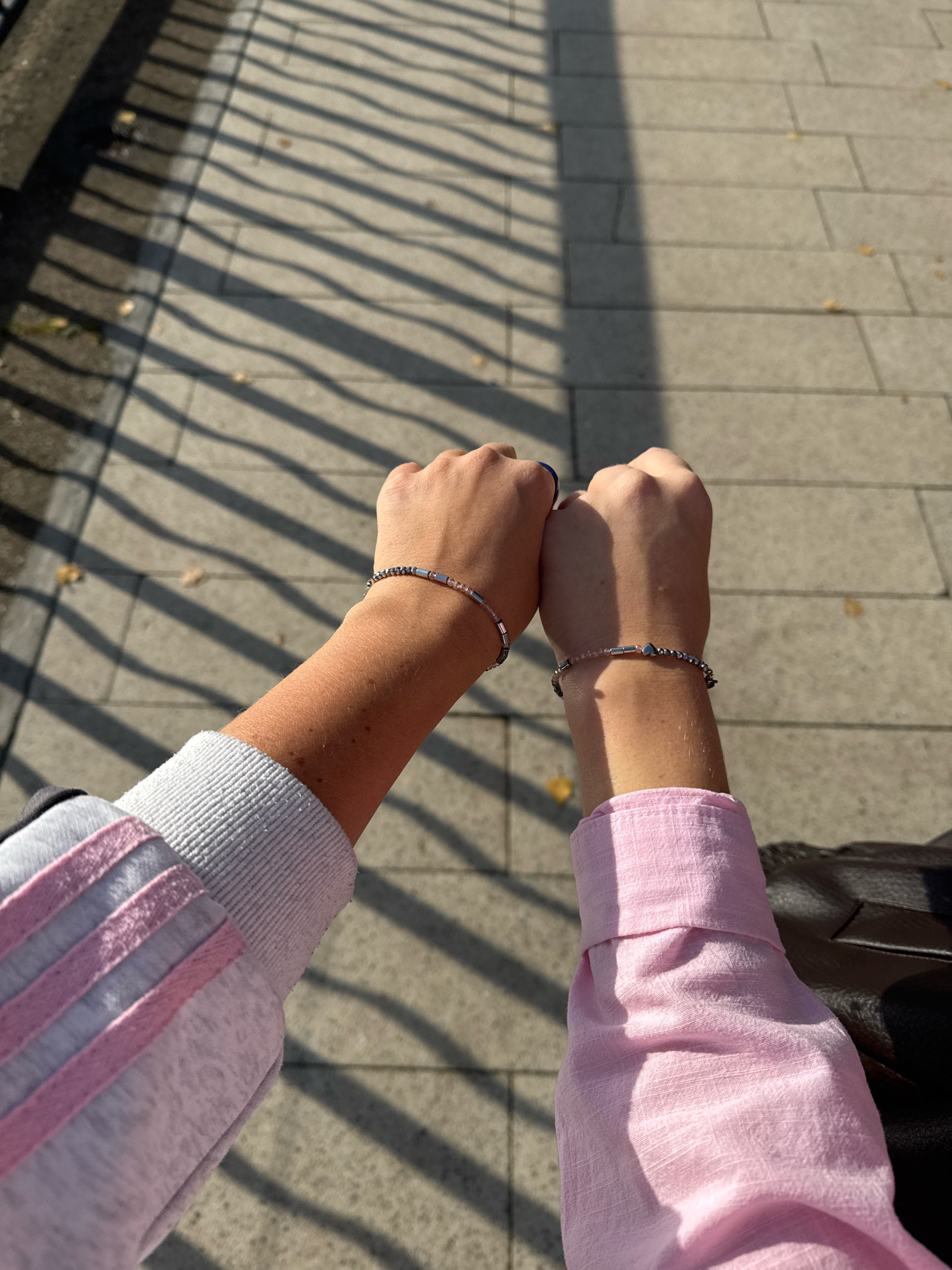 Two individuals, seen from the forearms down, stand with their fists gently touching. Both wear bracelets on their wrists. The person on the left has a light gray sweatshirt with pink stripes on the sleeve. The person on the right wears a light pink long-sleeved shirt. They are positioned outdoors on a paved walkway with a shadow pattern cast by a railing. The ground is composed of rectangular grey paving stones. The location is the city of Chaikovsky, Russia.