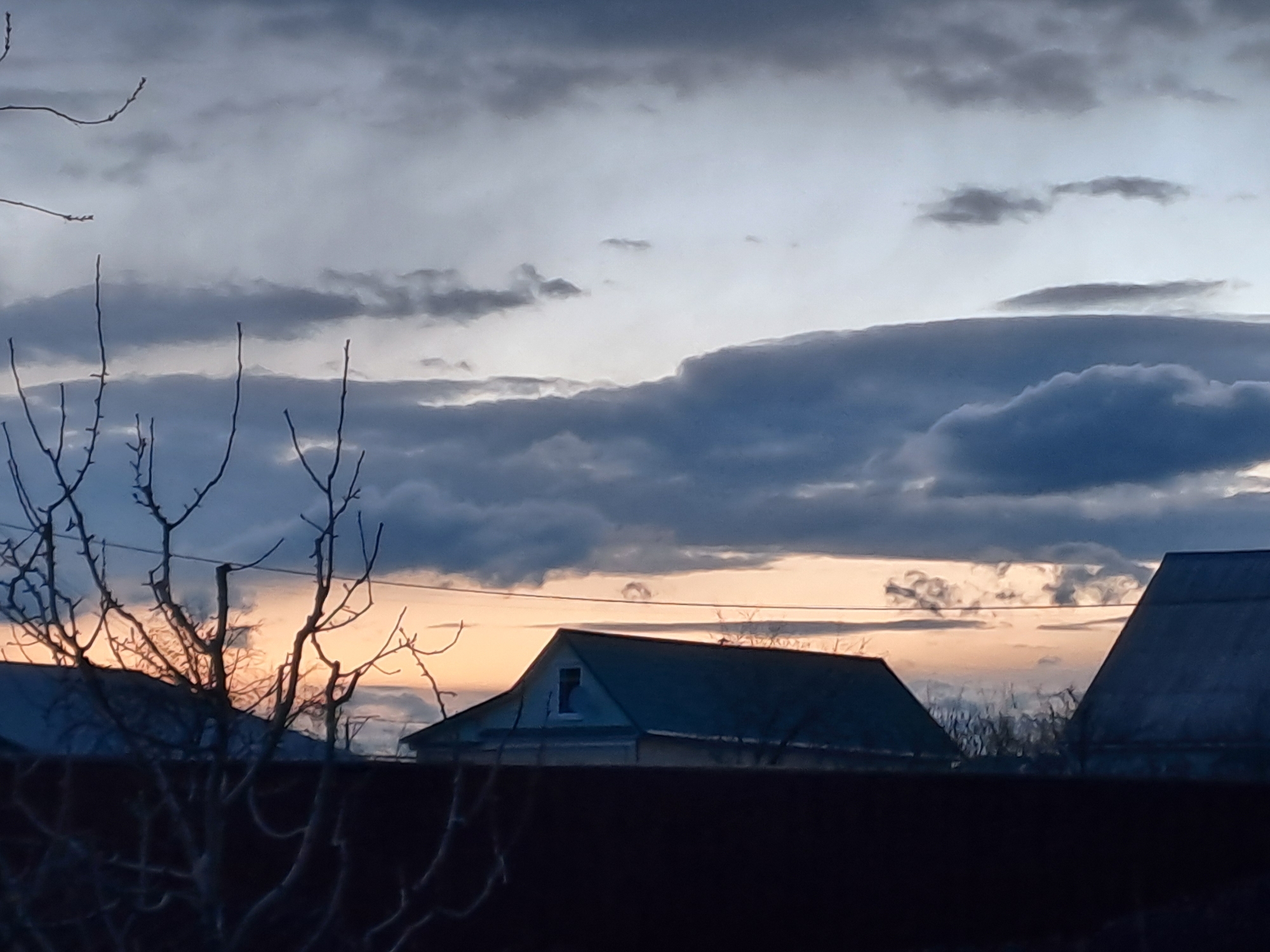 The image captures a tranquil rural or semi-rural landscape at either dawn or dusk, characterized by soft, transitioning light. The sky is a prominent feature, displaying a gradient from deep blues and dark greys in the upper regions to warm orange and light pink hues along the horizon, suggesting the sun's low position. Numerous clouds, ranging from heavy, dark formations to lighter, wispy streaks, fill the sky.
In the immediate foreground, a dark, solid fence or wall runs horizontally across the bottom of the frame. Above this, the intricate silhouettes of bare tree branches, devoid of leaves, dominate the left and central parts of the midground, indicating a season outside of summer. These branches create a natural, somewhat intricate foreground element.
Beyond the fence and trees, several house roofs are visible, largely in shadow. A prominent house, positioned centrally, features a dark, pitched roof with a small, visible dormer window. Portions of other roofs are also seen on the far right and left, suggesting a small settlement. The architecture appears typical of detached homes or dachas.
No people are visible in the scene, and no specific activities are taking place. The overall mood is peaceful and calm, with the dramatic sky providing a striking backdrop. There is no visible text within the image. The location context places this scene in Полянки, Russia.