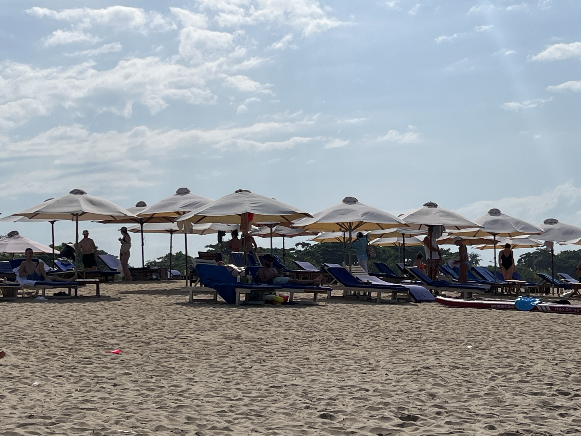 This image captures a wide view of a sandy beach in Nha Trang Ward, Vietnam, under a partly cloudy, light blue sky. The foreground and mid-ground are dominated by light brown sand with visible textures. A line of numerous light-colored beach umbrellas is evenly spaced across the mid-ground, providing shade for multiple blue sun loungers.
At least ten individuals are present within the frame. On the left, a male individual in blue shorts sits on a sun lounger. Towards the center-left, two male individuals stand, one wearing a hat and dark shorts. Another female individual is visible standing nearby. In the center-right, a male individual reclines on a blue sun lounger. Further right, a woman in a black one-piece swimsuit stands, and another person in a light blue top walks away from the viewer.
On the sand in the mid-right, a red paddleboard or surfboard with "seabeas" written on it is positioned horizontally, accompanied by a blue bag or inflatable item. A small pink, flat object is visible on the sand in the lower-middle foreground. In the background, behind the line of umbrellas, lush green foliage and trees are visible, indicating a vegetated coastal area. The scene is brightly illuminated, consistent with daytime.