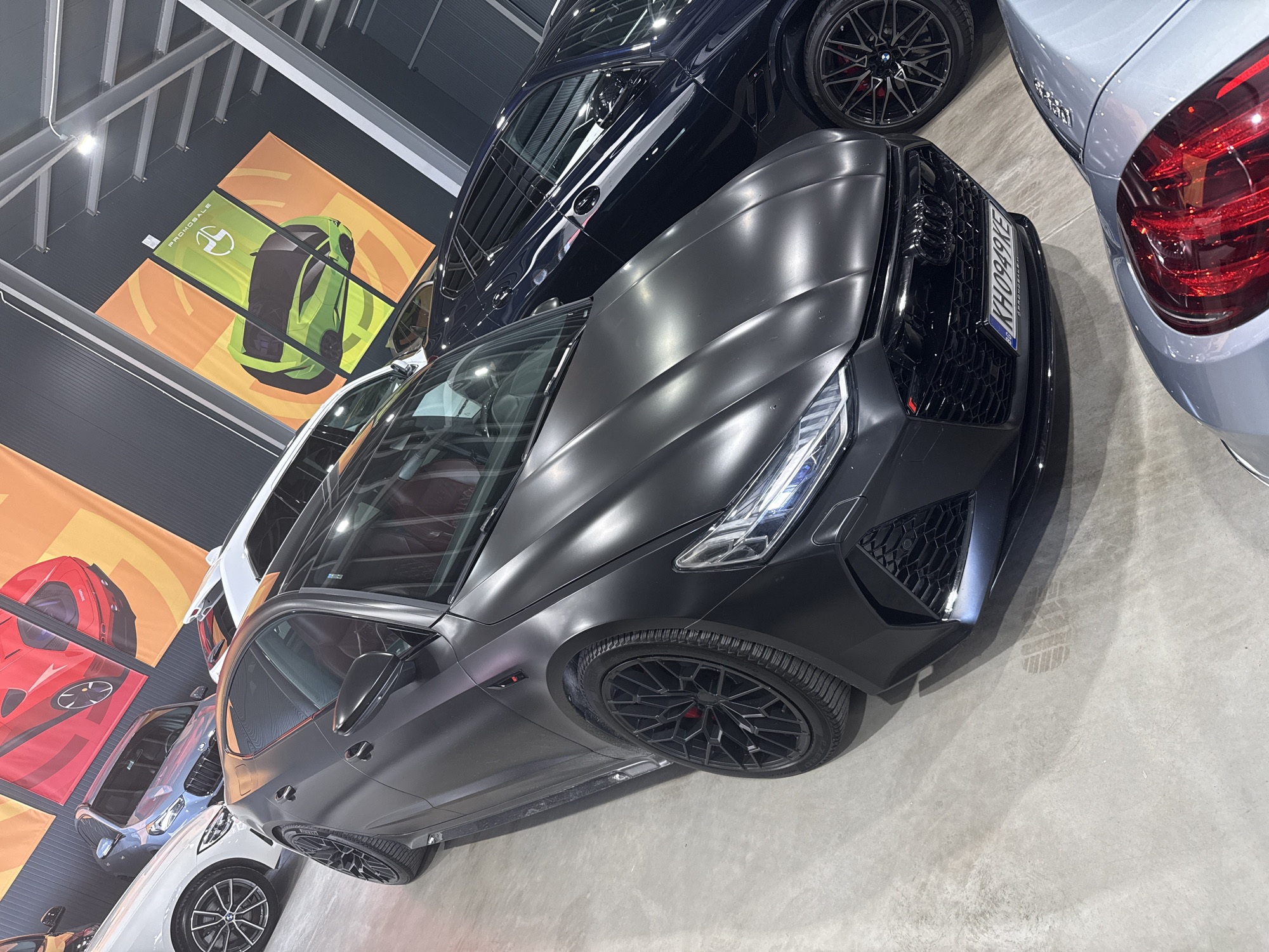The image depicts a car showroom interior, likely in Plovdiv, Bulgaria. Dominating the foreground is a matte black Audi RS6, featuring a black grille and rims. A blue BMW M5 is behind it, partially visible, and a light gray BMW M3 is seen on the right side, with its red taillight prominently displayed. In the background, orange and yellow banners with images of cars can be seen. The floor is a smooth, light gray concrete. The scene is well-lit with overhead lights visible at the top of the image. The environment suggests a commercial or promotional setting for automotive sales.