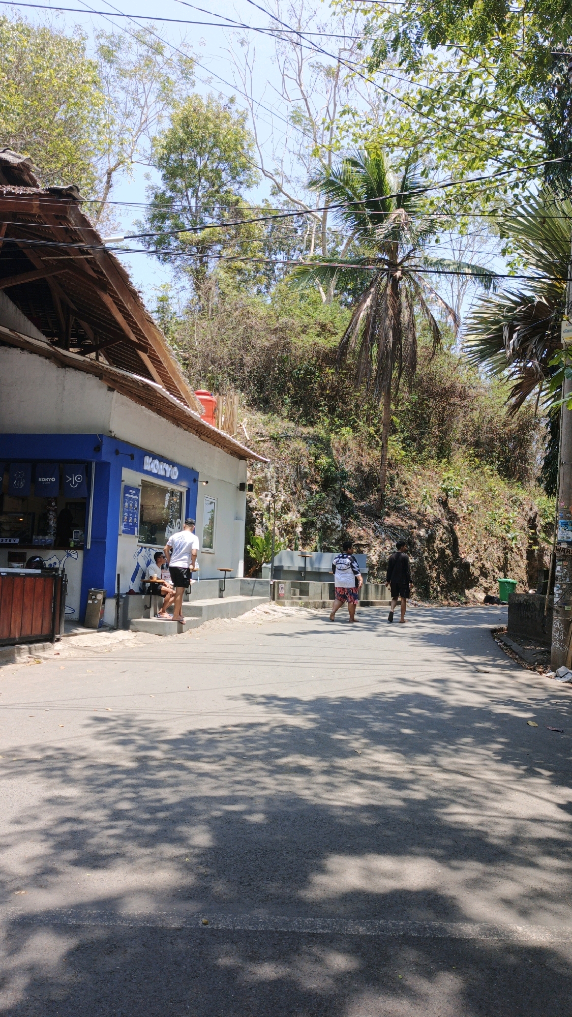 A street scene in Desa Batukaras, Indonesia, features a small, white building with a blue facade, possibly a cafe or shop, labeled "KOKYO" in prominent lettering. The building has a sloped, thatched roof and an outdoor seating area. Three people are visible outside the building: two young men are seated on steps, and one man stands near them. Further down the street, two individuals are walking away from the camera. The street is paved and dappled with shadows from overhead trees. The background consists of a steep, vegetated hillside and palm trees, indicative of a tropical environment. Overhead power lines are visible against a clear sky.