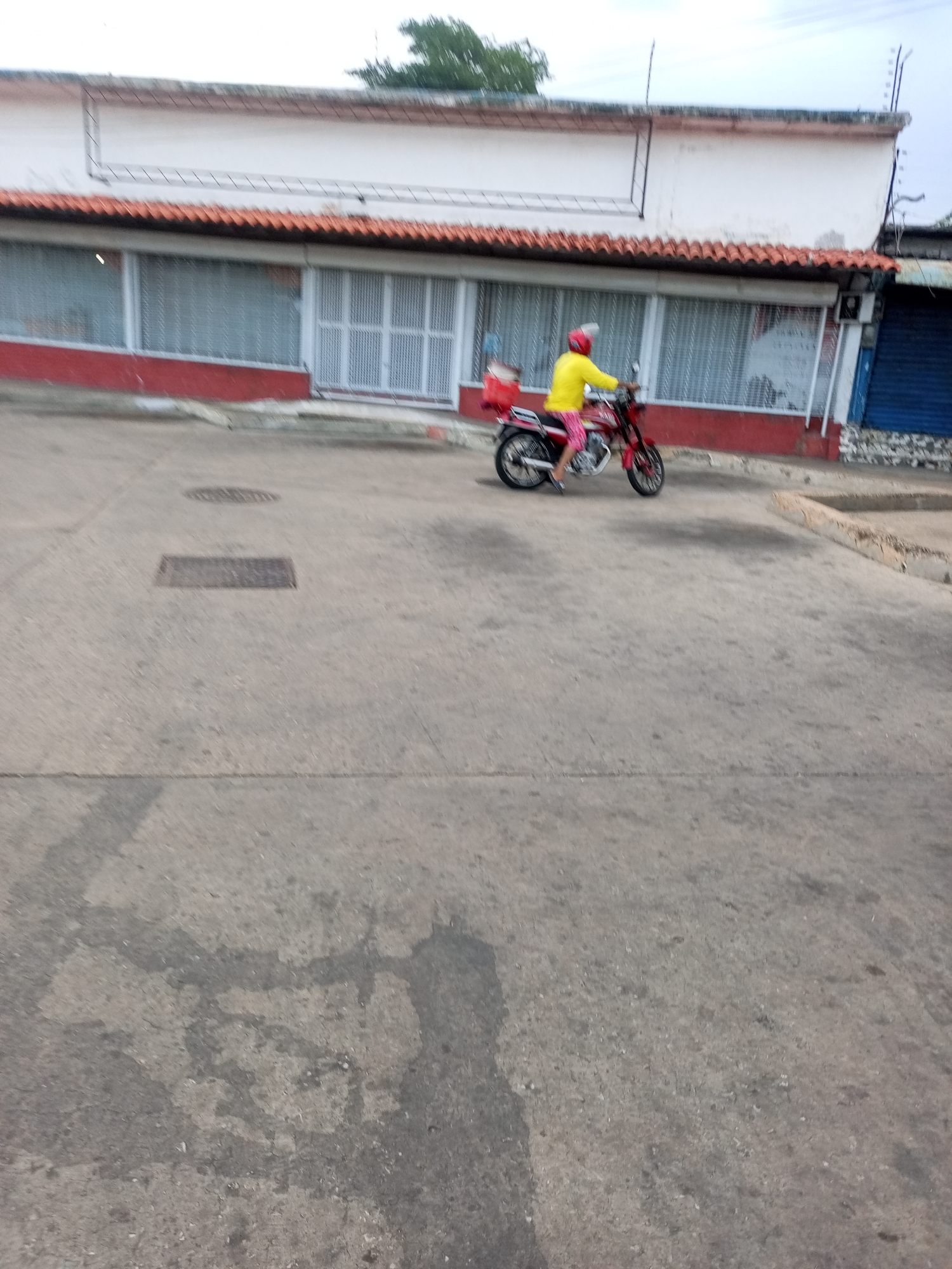 A person wearing a yellow shirt and pink shorts rides a red motorcycle with a red box on the back. The motorcycle is red with silver accents. The person is wearing a helmet with a clear visor. They are riding on a paved road in front of a white building with a red base and a tiled roof. The building has large windows with white shutters. The road is gray and cracked in places, with manhole covers visible. The sky is overcast. The scene appears to be in an urban or commercial area.