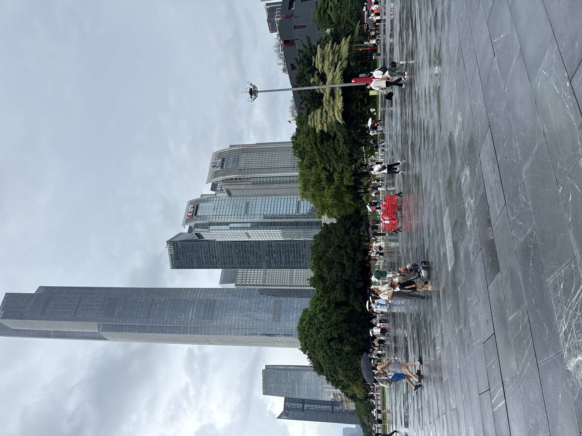 In Tianhe District, China, a rainy day is depicted with a wide, wet, grey stone plaza in the foreground. Numerous people are walking across the plaza, some holding umbrellas, indicating light rain. In the midground, a group of trees forms a dark green barrier, behind which various modern skyscrapers rise into a cloudy, overcast sky. Among the people, a few individuals are notable: two women walking with one holding a black umbrella, a woman pushing a child in a stroller, and several groups of people walking in different directions. Red barriers are visible on the left and right edges of the plaza, suggesting a cordoned-off area or construction. The architecture of the skyscrapers is diverse, with sleek glass facades and some featuring visible building names like "IMP".