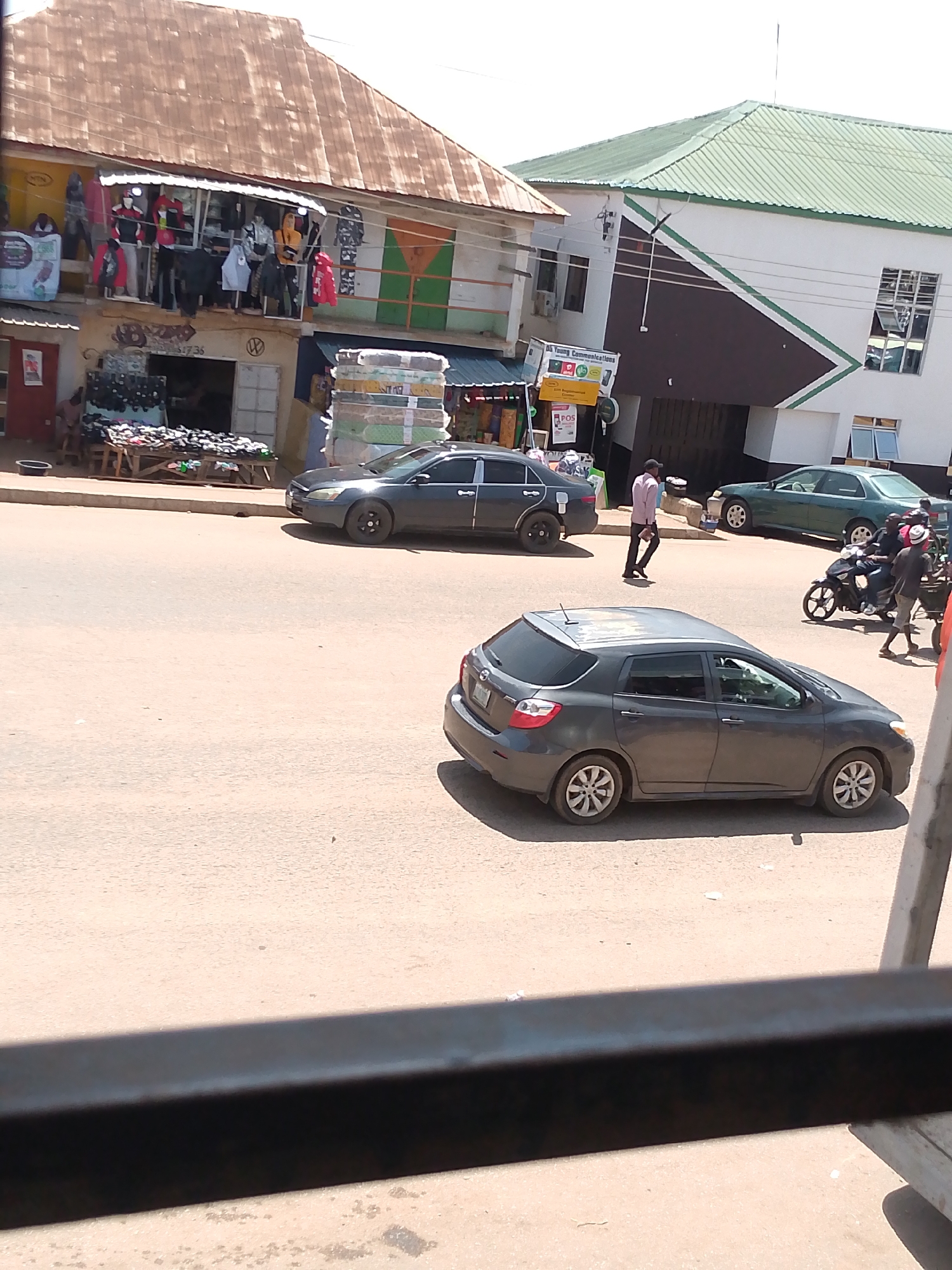 This image captures a street scene in Bukuru, Nigeria. In the foreground, a dark grey hatchback car is parked on the right side of the road. Further back, on the left side of the road, a dark grey sedan is in motion. In the background, a man in a pink shirt and dark pants is walking on the sidewalk. A group of people are on a motorcycle, with one person driving and another person sitting behind them. The buildings lining the street have a mix of architectural styles. One building on the left has a corrugated iron roof and a storefront displaying clothing on mannequins. Another building on the right is painted white with a green roof and features a prominent diagonal design in brown and green. The street is paved with asphalt, and the overall environment appears to be an urban setting.