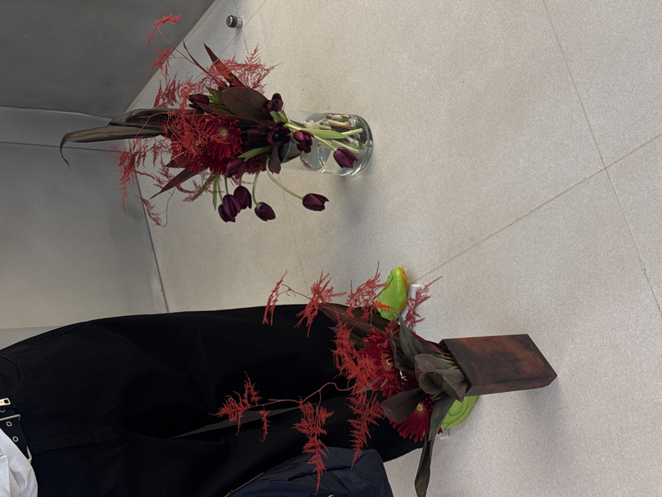 Two floral arrangements sit on a gray tiled floor. One arrangement is in a clear glass vase with dark purple tulips and red flowers. The second arrangement is in a dark brown wood-like container with red flowers and dark green leaves. A person wearing black pants and neon green shoes is present near the second floral arrangement. Part of a white shirt is visible at the waist, and a blue jacket is lying in the lap.