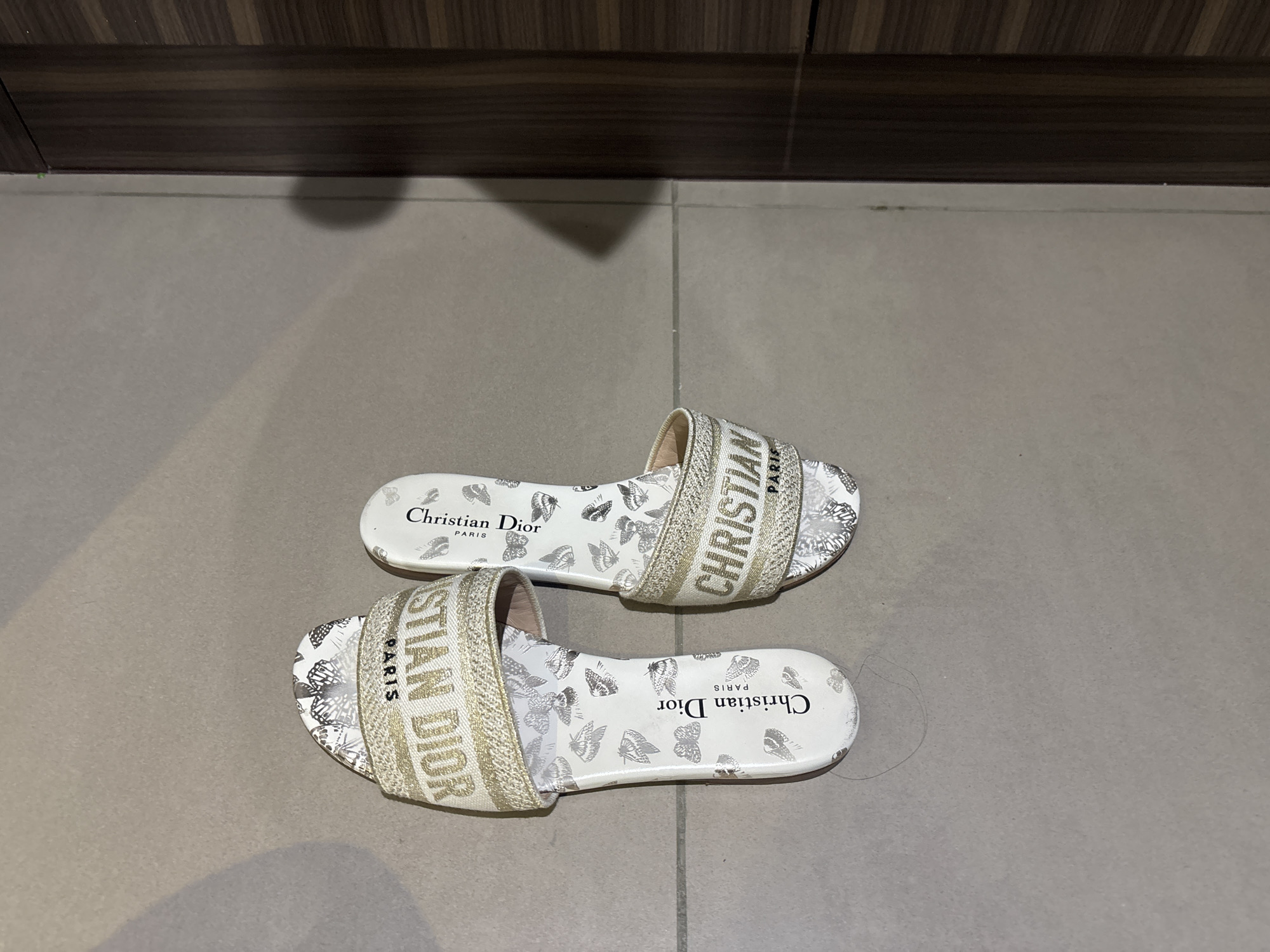 A pair of white Christian Dior slide sandals with a butterfly pattern and gold lettering is displayed on a tiled floor. The sandals are positioned side-by-side, with the left sandal slightly angled forward. The right sandal is positioned slightly behind and to the right of the left sandal. The floor tiles are a light grey, and the background to the right features a dark wood cabinet or door. The lighting suggests an indoor environment. There is a faint shadow cast on the wall to the right, possibly from the cabinet. The scene is devoid of people and any specific indicators of its location within Dubai, United Arab Emirates.