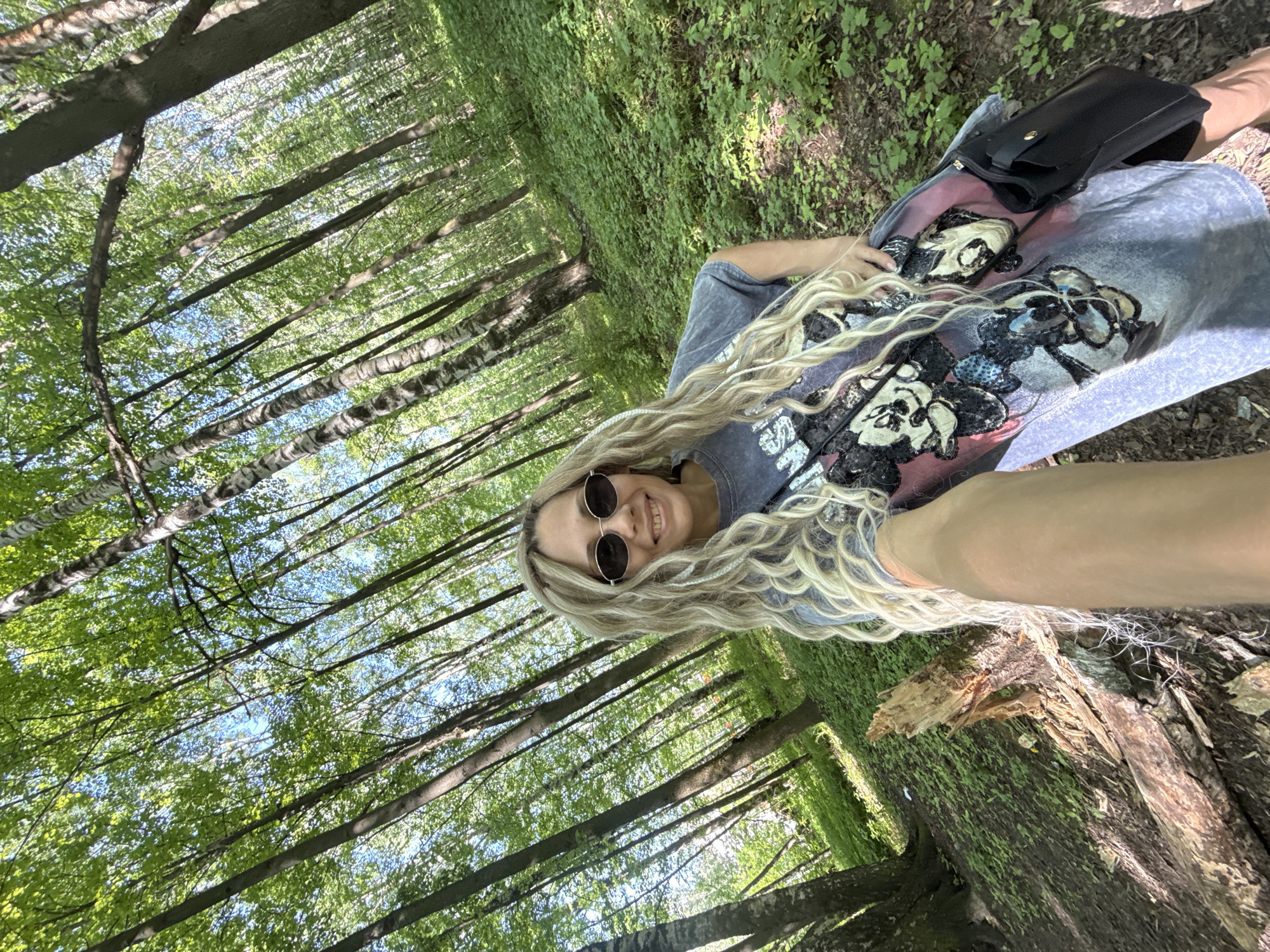 A young woman is lying on a tree trunk in a forest. The forest consists of tall, thin trees, with green foliage visible in the canopy. The woman is wearing a printed t-shirt, sunglasses, and has long, blonde, wavy hair. She is also carrying a black purse. The location is in an unknown city. The environment suggests a natural, wooded area.