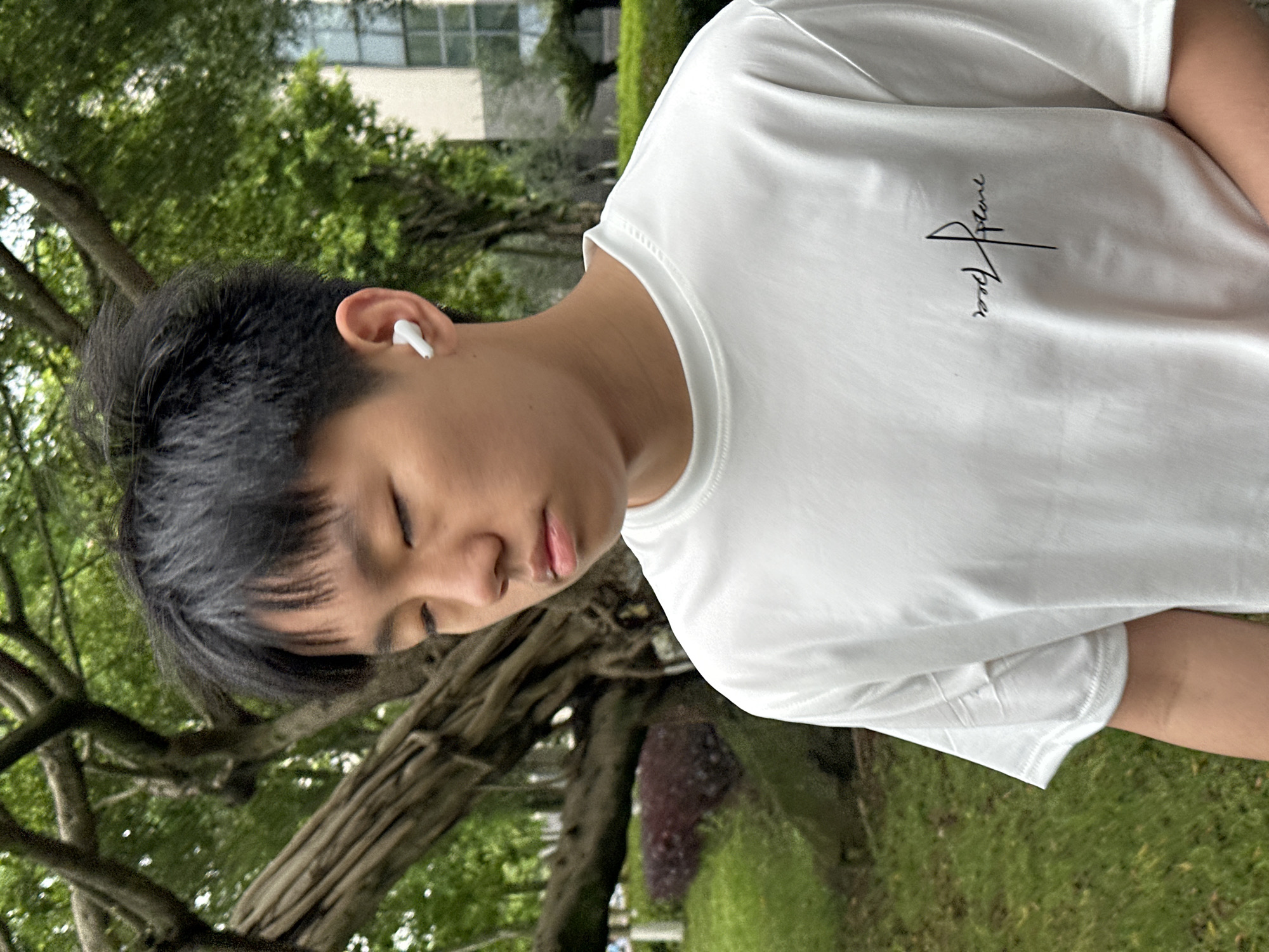 A young Asian male wearing a white t-shirt is outdoors. He has short, dark hair and an earbud in his left ear. His eyes are closed. The background features trees and grass, indicating a park or green space. Buildings are visible in the distance. The location is Jiangjin, China.