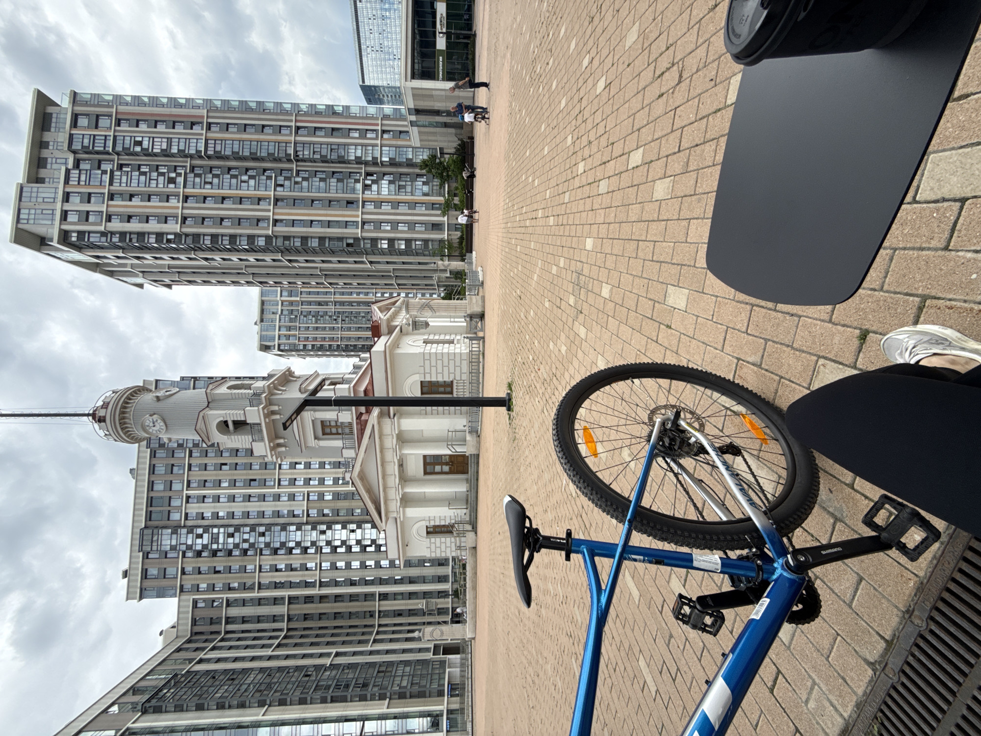 The image depicts an urban scene in Moscow, Russia. A blue bicycle lies on a brick-paved surface, with a black laptop bag nearby. In the background, modern high-rise buildings frame a classical white building with a clock tower. Several people are walking on the brick path. The sky is overcast.