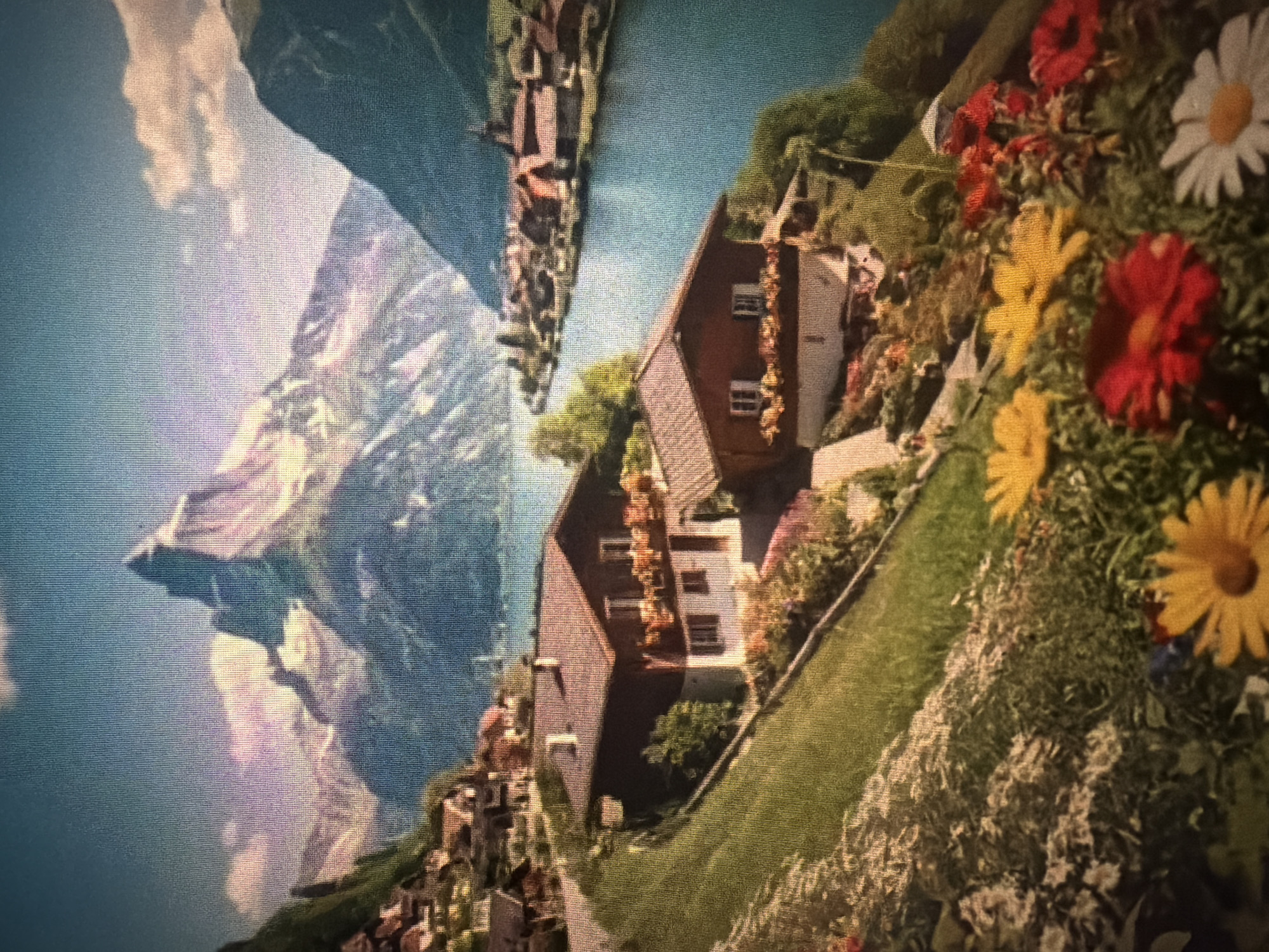 The image depicts a scenic mountain landscape with a lake and a village. In the foreground, there are several houses with traditional alpine architecture, nestled on a green hillside adorned with colorful flowers. The houses have brown roofs, wooden balconies with flower boxes, and white-trimmed windows. A winding path leads through the houses, and a grassy slope with more flowers is visible.
In the mid-ground, a bright blue lake stretches out, reflecting the surrounding mountains and sky. Along the far shore of the lake, a charming village with similar architectural styles is visible, appearing to be built into the hillside.
The background is dominated by majestic, snow-capped mountains under a clear blue sky with scattered clouds. The mountain peaks are sharp and rugged, conveying a sense of grandeur.
The image does not depict the city of Baan Mae Nam, Thailand. Instead, it portrays a European alpine setting, likely in Switzerland or a similar mountainous region known for its lakes and villages. There are no people or animals visible in the scene. The overall atmosphere is peaceful and picturesque.