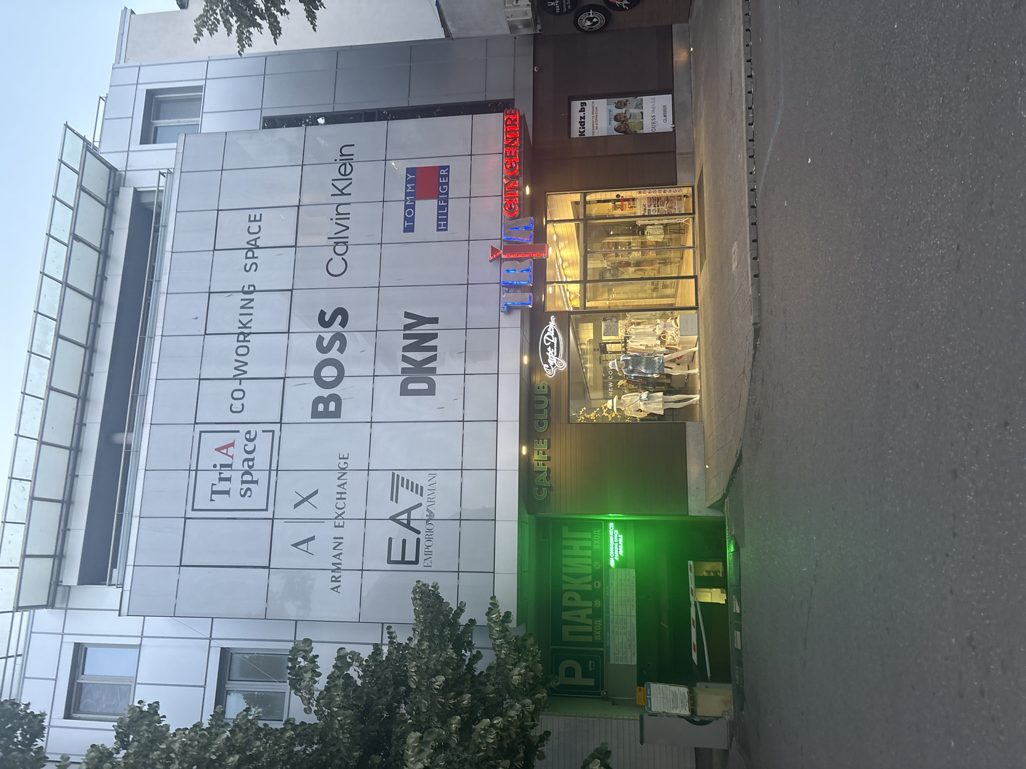 The image shows the exterior of the TRI A CITY CENTRE building in Burgas, Bulgaria. The building has a modern facade with various brand logos displayed on the upper levels, including Armani Exchange, Emporio Armani, BOSS, Calvin Klein, DKNY, and Tommy Hilfiger. The ground floor features shop entrances and a parking garage entrance. The parking garage entrance is illuminated with green lights, and a barrier gate is visible. A "CAFFE CLUB" sign is also present on the ground floor. Mannequins are displayed in the window of a shop. The overall scene depicts a commercial building in an urban setting, likely during daylight hours.