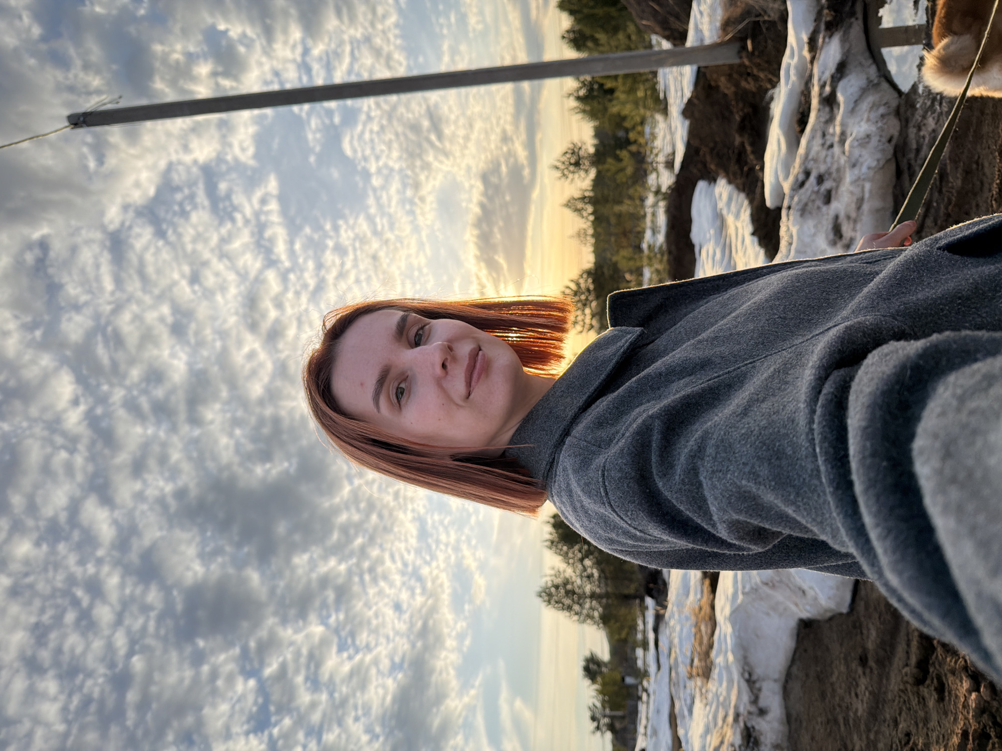 The image is a rotated selfie of a woman with short auburn hair. She is wearing a gray coat. Behind her is a landscape with snow, trees, and a sky filled with clouds. The sun is setting, casting a golden glow. A metal pole is visible on the left side of the image. The location appears to be in a rural or semi-rural area, potentially near the city of Chaikovsky, Russia. A leash is visible.