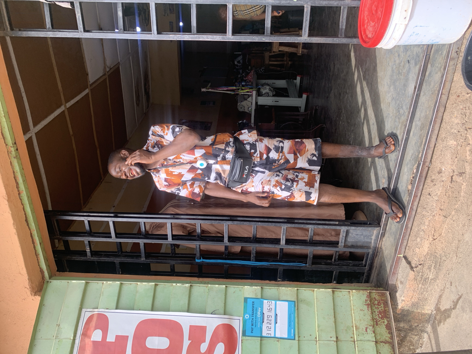The image shows a man standing in the doorway of a business in Jos, Nigeria. The man is wearing a patterned shirt and shorts, and a Fila fanny pack. He is smiling and appears to be talking on a cell phone. Inside the building, there is a sewing machine and other equipment. A sign visible reads "PAY WITH TRANSFER" and lists an OPay account number: 3 12229 1643. The exterior of the building appears weathered.