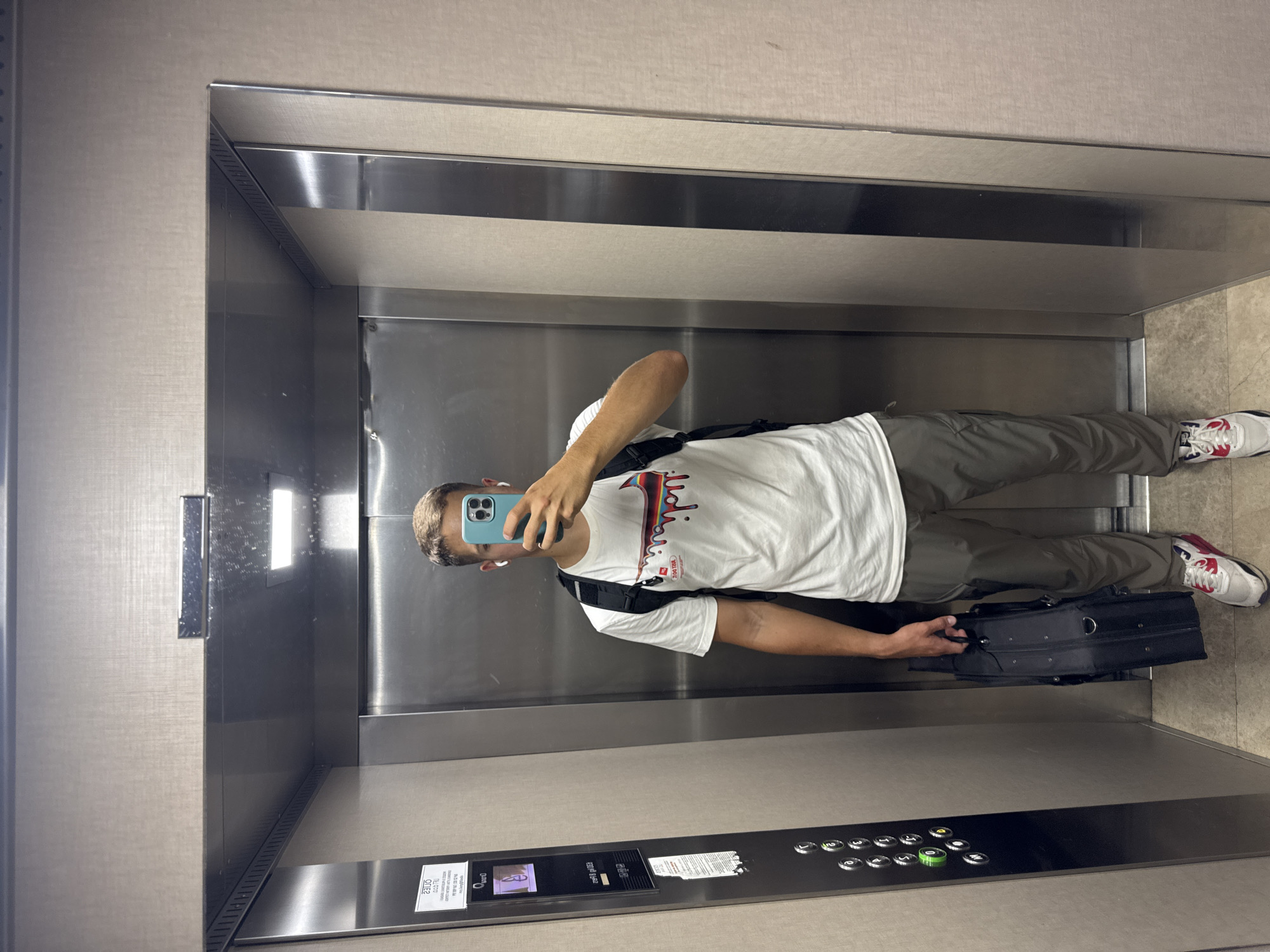 In Budva, Montenegro, a young man stands in an elevator, holding a black briefcase in his right hand. He is wearing a white t-shirt with a colorful graphic design, grey cargo pants, and athletic shoes. He is also wearing white wireless earbuds and has a black backpack on his shoulders. The elevator has brushed metal walls and a textured ceiling. A panel with buttons and a small screen is visible on the left side of the elevator. The man is using his phone to take a selfie.