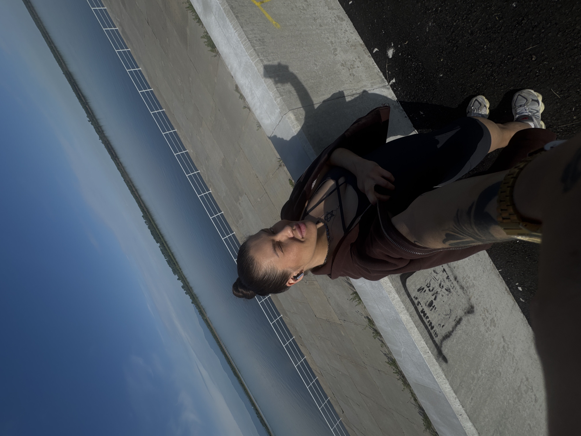 In a selfie taken outdoors, a woman with her hair pulled into a bun is captured at a slight angle. She is wearing a dark top with a harness-like detail and a brown zip-up hoodie. In her right ear, she has a wireless earbud. The background features a wide body of water, possibly a river or lake, bordered by a tree line and distant mountains under a clear blue sky with some wispy clouds. A white railing runs along a concrete structure next to the water, and the woman is standing on a paved area adjacent to a concrete wall. Graffiti is visible on the concrete surface in the foreground. The camera angle suggests the photo was taken from a raised vantage point. The woman is looking towards the camera with a slightly closed eye and an open mouth, and the shadow of a person holding a phone is cast on the wall.