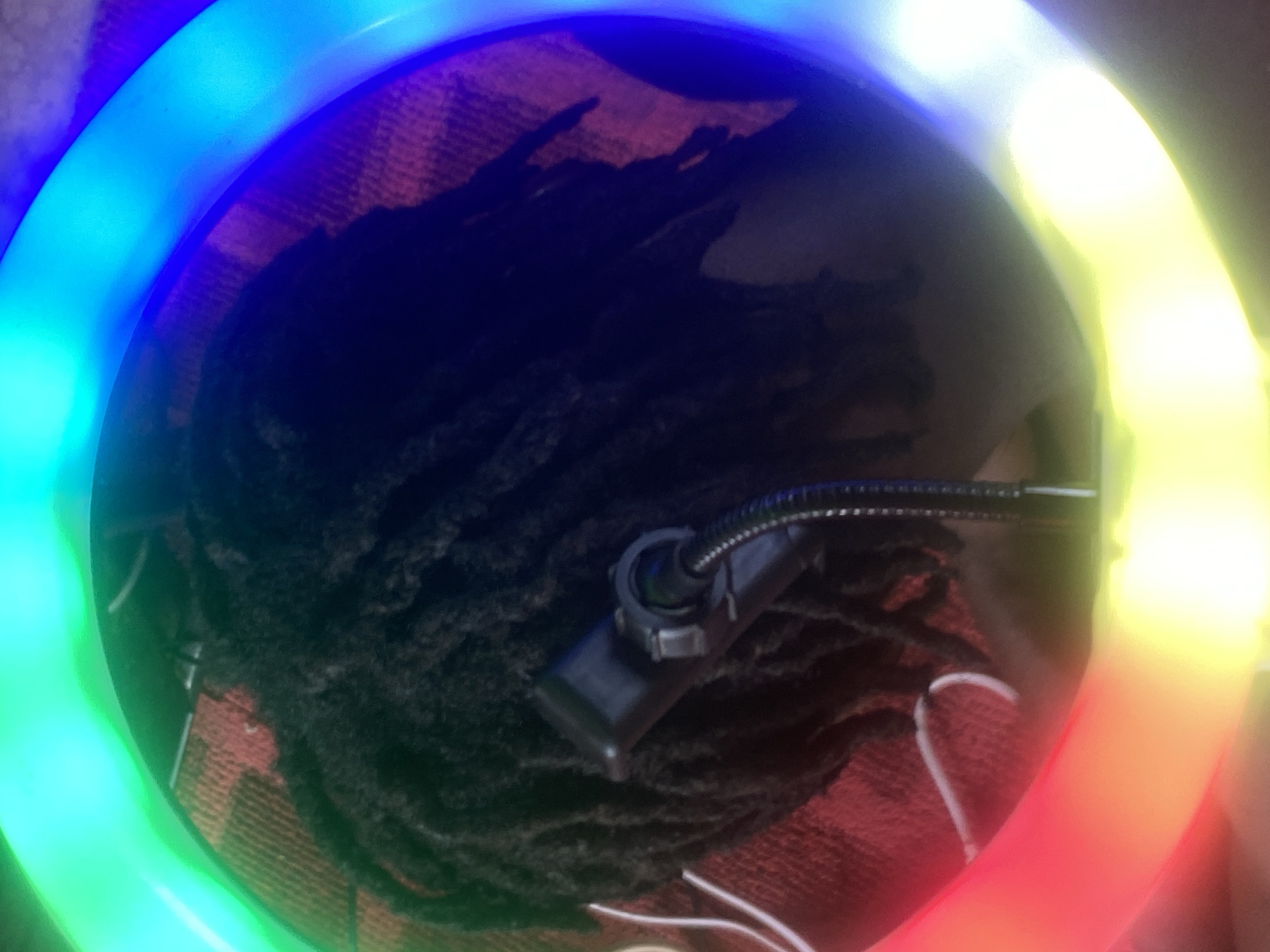 The image shows a close-up view of a person's head from above, seemingly positioned within the ring of a multicolored LED ring light. The person has dark hair styled in dreadlocks. At the base of the neck, what appears to be a plaid fabric or clothing is visible. There is a black mounting bracket attached to the ring light. The location is not discernible from the image.