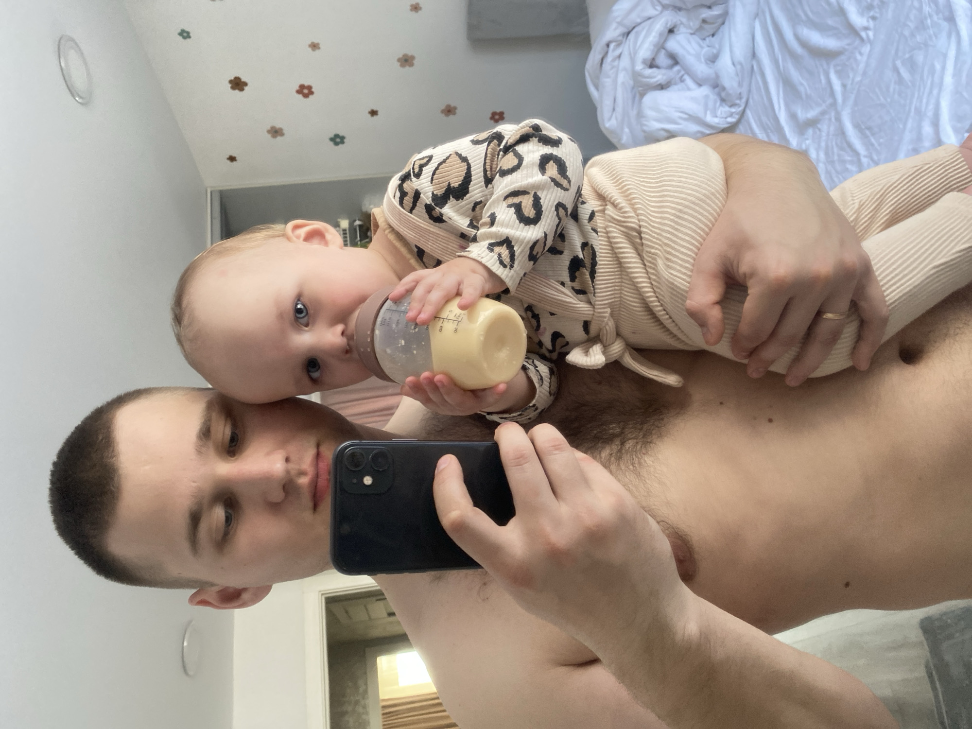 A shirtless man with short-cropped hair holds a baby wearing a leopard-print top and beige pants in his arms, while taking a selfie with a black smartphone. The baby is holding and drinking from a baby bottle. The location appears to be inside a home or apartment, based on the interior details like wall decorations and a bed with white rumpled bedding visible in the background. No evidence is present to indicate the city of Chaikovsky, Russia.