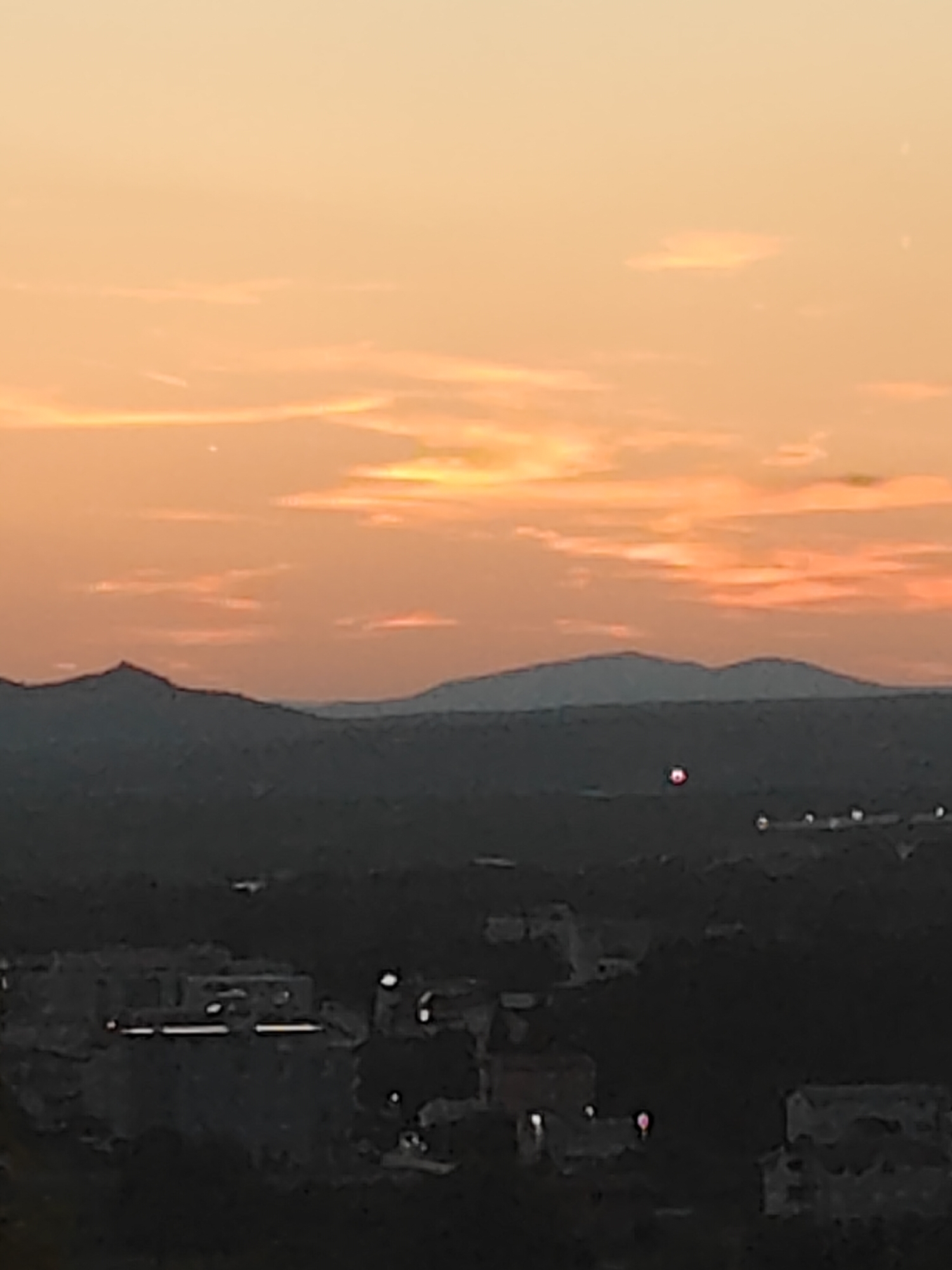 This is a long-range, low-resolution photograph of a landscape at dusk, likely taken from an elevated position overlooking a town. The sky is a gradient of warm orange and yellow hues, with thin wispy clouds scattered across it. In the mid-ground, silhouetted mountains form a dark, jagged horizon line. Below the mountains, the landscape transitions into a darker, indistinct area that appears to be a town or city. Numerous small, bright lights are visible in this area, suggesting artificial illumination from buildings or streetlights. In the center of the image, a small, bright red circular object is visible in the sky, and several other smaller, white lights are scattered throughout the darker landscape. The image quality is poor, with significant noise and blur, making detailed identification of specific structures or objects challenging. The scene depicts a rural or semi-rural setting at twilight, with a prominent natural landscape element (mountains) dominating the background and a populated area in the foreground.