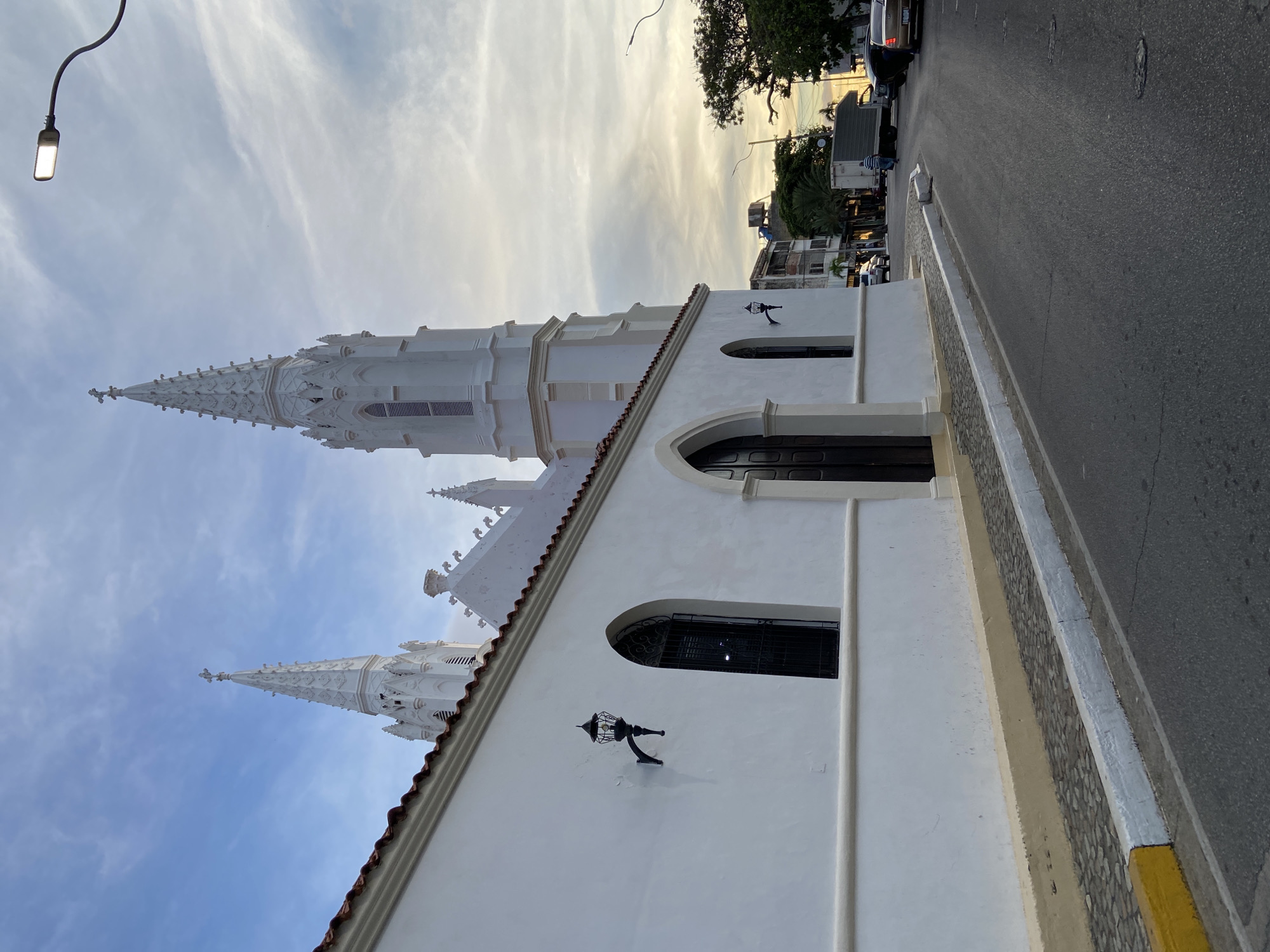 The image depicts the Iglesia de Nuestra Señora de la Asunción in Juan Griego, Venezuela, bathed in the soft light of a cloudy dusk. The stark white facade of the church dominates the left side of the frame, its architectural details, including arched windows and decorative spires topped with crosses, standing out against the pale sky. Ornate black lanterns are mounted on the wall. A cobbled pedestrian walkway runs along the base of the church, bordered by a yellow and white curb. The street, paved with asphalt, curves away from the church into the distance, where other buildings and trees are visible. A few vehicles, including a white truck and several cars, are present on the street. The sky is a canvas of muted blues, grays, and yellows, with wispy clouds indicating the setting sun. The overall scene suggests a quiet evening in a coastal town.