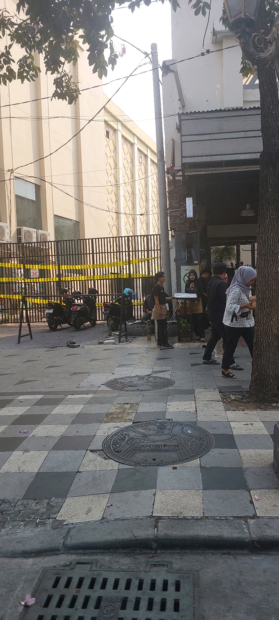 The image depicts a street scene in Surabaya, Indonesia, focusing on a sidewalk area adjacent to a building with a dark awning. In the foreground, the sidewalk is paved with a checkered pattern of grey and beige square tiles. Two circular manhole covers with intricate designs are visible on the pavement. A drain grate is also present at the lower edge of the image.
Several people are present on the sidewalk. One individual wearing a grey and white checkered top and a headscarf is walking away from the camera, holding a brown paper bag. To their left, a group of individuals, some wearing aprons, appear to be gathered near the entrance of the building. One person in the group is holding a silver tray. Behind them, further to the left, is a black metal fence with yellow caution tape stretched across it. Several motorcycles are parked behind the fence.
The building in the background has a light beige facade with a repeating diamond pattern and visible air conditioning units. A tree with visible bark stands to the right of the image, partially obscuring the building. Power lines are strung across the scene, and a utility pole is visible. The overall impression is of a public urban space with commercial activity.