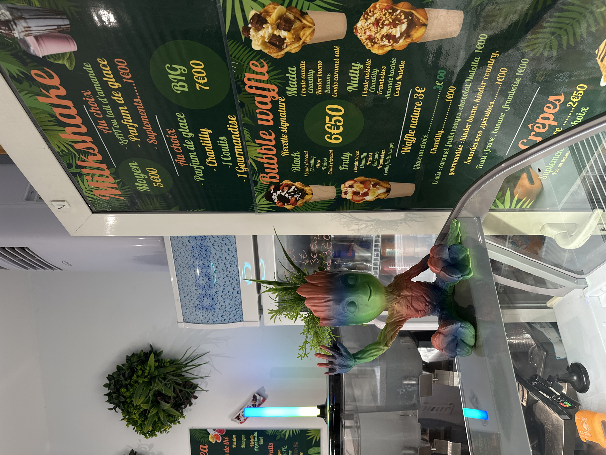 The image shows a close-up of a menu board in a food establishment. The menu board features colorful graphics and text advertising various food items like milkshakes, bubble waffles, and crepes. There are images of the food items on display, enticing customers. In the foreground, a small, rainbow-colored figurine of Groot from Guardians of the Galaxy is placed on the counter, adding a playful touch to the scene. The background includes elements of the establishment's interior, such as refrigeration units and other display cases, suggesting it's a shop or cafe. The overall lighting and composition create a vibrant and inviting atmosphere.