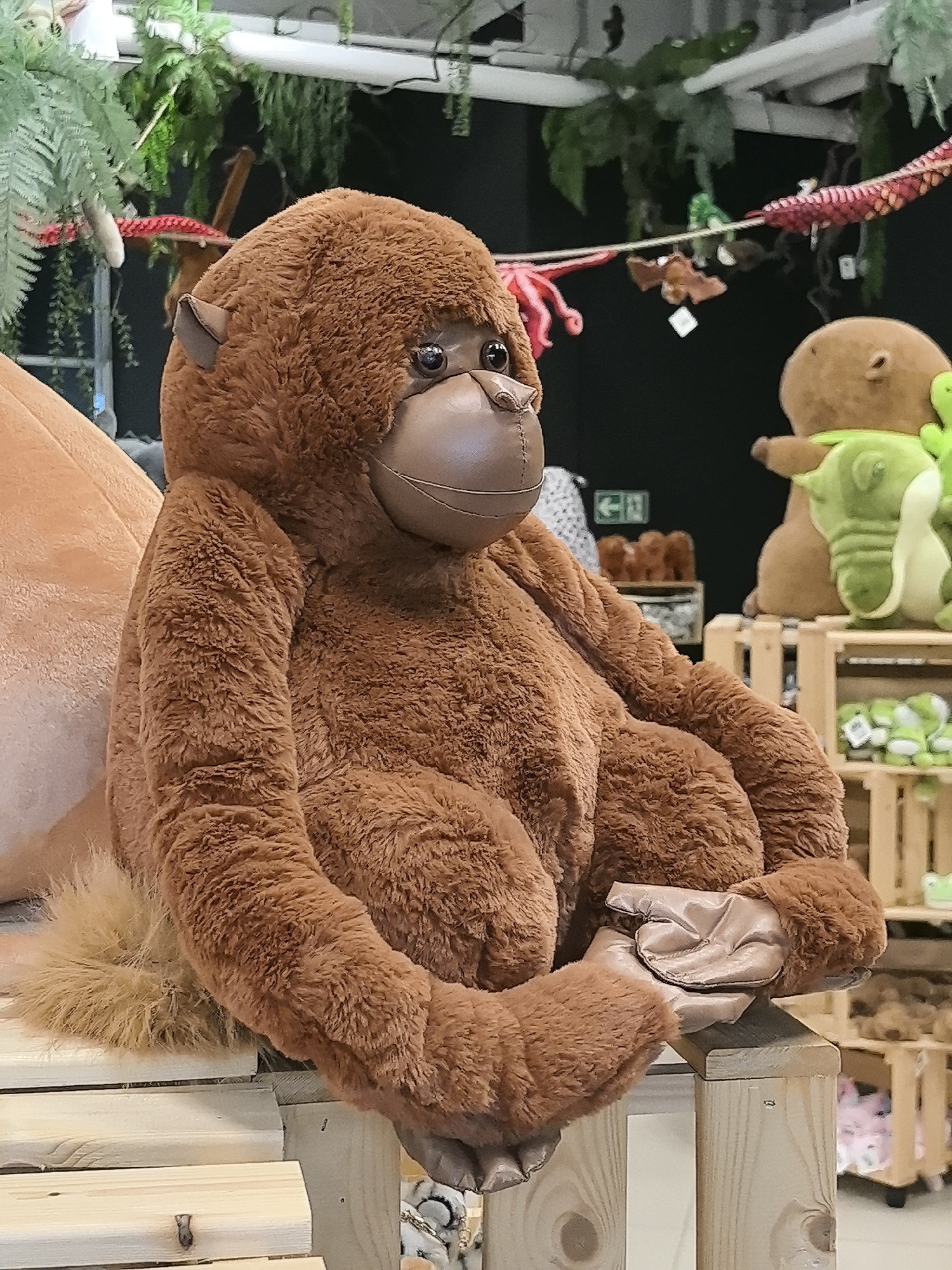 A large, plush, brown, stuffed orangutan toy sits on a wooden structure. The orangutan is posed in a seated position with its arms and hands placed in front of its body, holding a brown object. In the background, other stuffed animals are visible, including a brown hippopotamus-like figure and a green crocodile. Wooden crates and greenery add to the scene. A sign with a green arrow is visible in the background. Based on prior knowledge, this image may be from a shop in Perm, Russia.