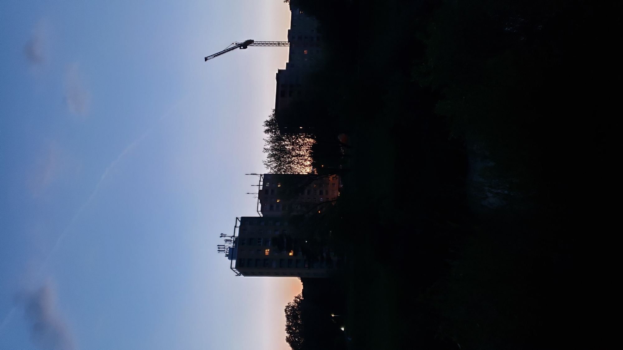 The image depicts a twilight scene in Prešov, Slovakia, with the sky transitioning from a deep blue at the top to a soft orange and purple hue near the horizon. Silhouetted against the sky are several high-rise apartment buildings and trees.
The most prominent building on the left is a tall, rectangular structure with multiple antennas on its roof. Several windows on its facade are illuminated, suggesting occupancy. Adjacent to it, slightly to the right and shorter, is another similar apartment building.
To the right of the apartment buildings, a construction crane stands tall, its framework stark against the sky. The silhouette of trees forms a dark band across the middle ground, with the setting sun or rising moon partially visible through their branches, casting an orange glow.
The foreground is dominated by a very dark, underexposed area, likely representing a park or a natural landscape at night, with minimal detail visible. The overall impression is one of urban development set against a tranquil evening sky. There are no people or discernible human activity visible in the frame.