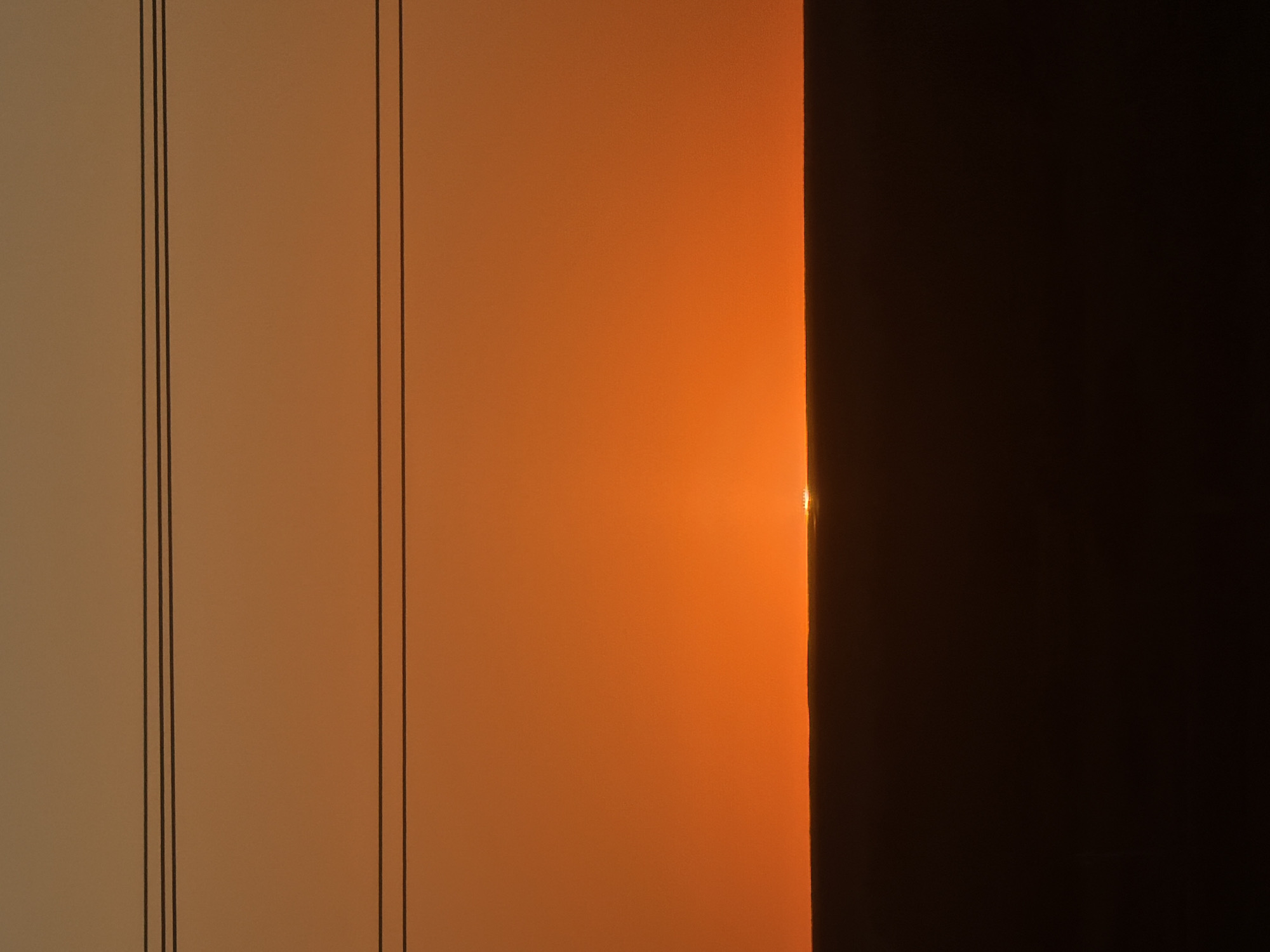 The image captures a sunset over a dark, flat terrain in Karamysh, Russia. The sky transitions from a deep orange near the horizon to a lighter, muted orange and then a pale beige at the top. Three horizontal power lines are visible across the upper portion of the frame. The sun is just a sliver of bright light peeking over the dark horizon line, suggesting it is either setting or rising. The foreground is a solid mass of dark brown, representing the ground or landscape. There are no people or animals visible in the scene. The overall impression is one of a stark and minimalist landscape at dusk or dawn, with human infrastructure hinted at by the power lines.