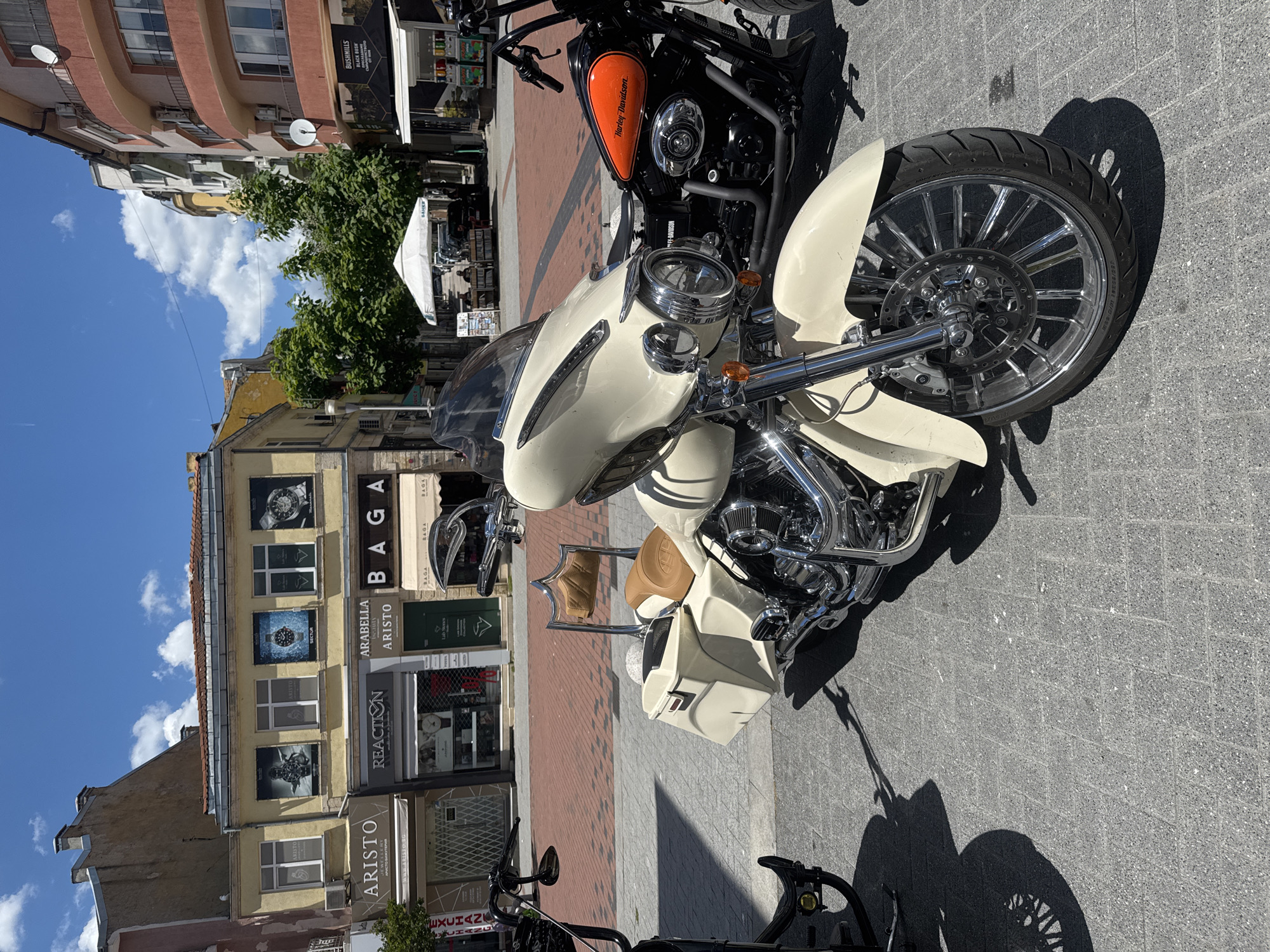 The image depicts a street scene in Varna, Bulgaria, specifically on Preslav Street, near the "Aristo" jewelry store, as indicated by the signage. The foreground features two parked motorcycles. The larger motorcycle in the center is a cream-colored cruiser with chrome accents, a large windshield, and saddlebags. To its right is a black and orange Harley-Davidson motorcycle. The background shows a commercial street with several shops, including "Aristo," "Reaction," and "Arabella Aristo." A white canopy tent is visible further down the street, suggesting an outdoor market or seating area. The sky is blue with scattered clouds, indicating a bright, sunny day. No people are clearly visible interacting with the motorcycles or the environment.