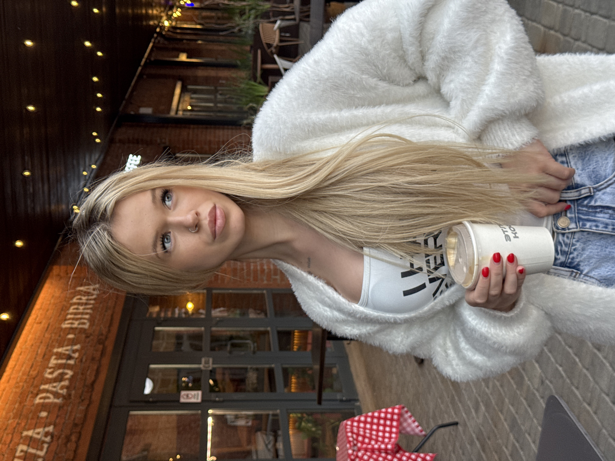 This is a portrait of a young woman with long blonde hair, holding a white paper cup with a clear lid. She is wearing a fluffy white cardigan over a white crop top and blue denim jeans. Her nails are painted red. She has a nose ring and is looking towards the camera with a slight smile. The image appears to be taken outdoors, with a brick building behind her that has the words "PIZZA. PASTA. BIRRA." painted on it. String lights are visible above and to the left of the woman, suggesting it might be evening or a dimly lit area. A red and white checkered tablecloth is visible in the lower right corner of the image, hinting at a dining or cafe setting. The overall mood is casual and relaxed. The text on the cup is partially visible and appears to be "STILL KOT."