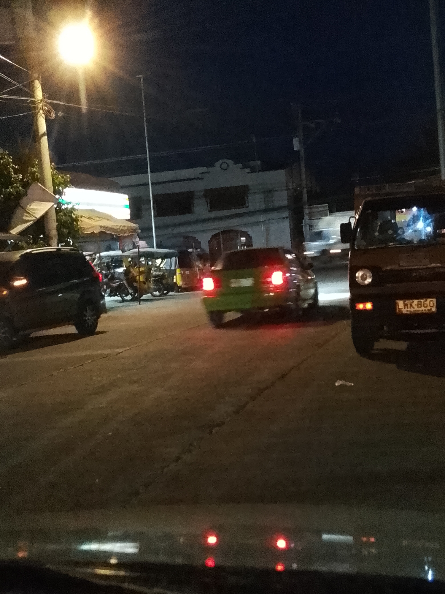 This media file depicts a street scene at night in Davao City, Philippines. The foreground is dominated by the interior of a vehicle, likely a car, as suggested by the dashboard lights and the view through the windshield. The road ahead is illuminated by streetlights and vehicle headlights, with a bright, diffused streetlight prominently visible in the upper left corner.
The scene shows traffic on a paved road. Several vehicles are present, including a dark SUV on the left, a green car with its brake lights illuminated in the center, and a dark-colored utility vehicle or van on the right with a license plate visible. Behind these vehicles, a group of three-wheeled motorized vehicles, possibly tricycles, are parked or moving slowly. The presence of these tricycles is a common sight in Davao City.
The background reveals a building with a light-colored facade and dark window openings. Another building, partially obscured, can be seen further down the street. The overall atmosphere is that of a typical urban environment at night, with the glow of artificial lights against the dark sky. The time of day is clearly night, and the weather appears to be clear.
The license plate on the utility vehicle on the right reads "LHK 860". The image captures a moment of transit, with vehicles navigating the road amidst the ambient city lights. There are no clear indications of specific events or strong emotions conveyed, but the scene suggests normal evening activity.