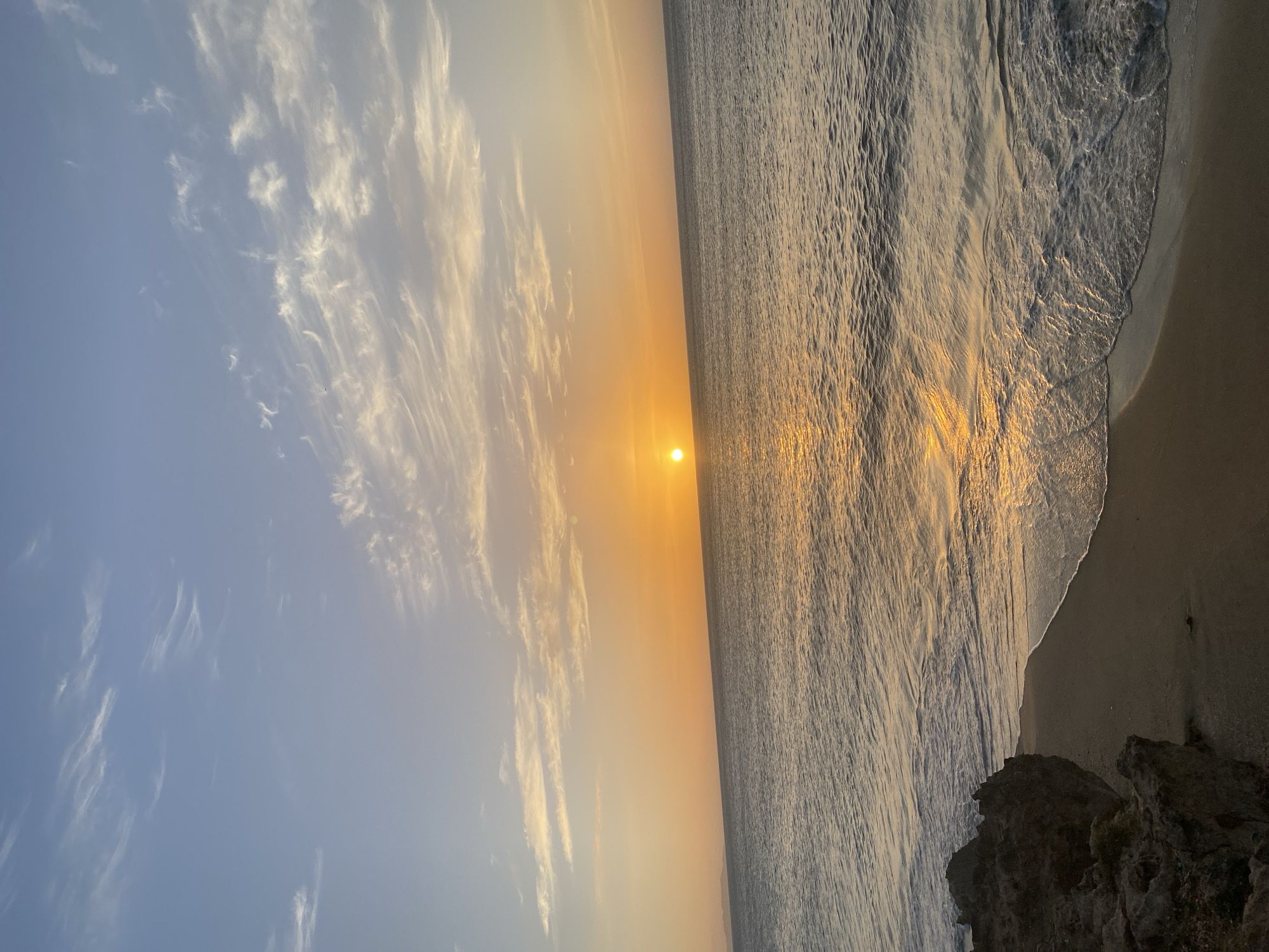 The image depicts a serene sunset over the Caribbean Sea, as viewed from the coastline of Juan Griego, Venezuela. The sun, a bright, warm orb, is positioned on the horizon, casting a golden, shimmering reflection across the rippling water. Wispy, light-colored clouds are scattered across a clear, pale blue sky that transitions to a soft orange hue near the horizon. The foreground features a dark, rocky outcrop on the left, meeting a sandy beach where gentle waves are washing ashore, creating frothy white foam. The overall scene conveys a sense of tranquility and natural beauty.