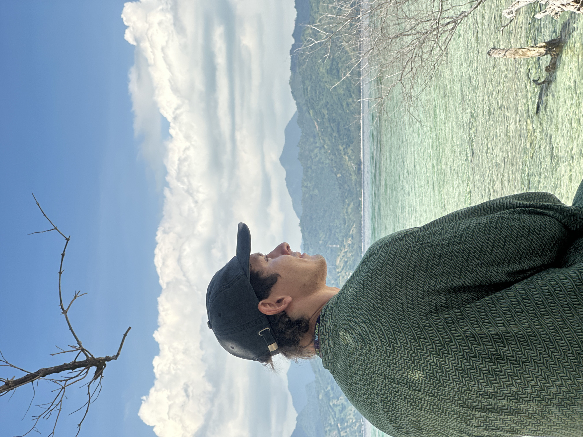 A vertically-oriented medium shot shows a man in the foreground gazing up into a sky filled with large, white cumulus clouds. He is wearing a black baseball cap and a dark green jacket with a textured herringbone pattern. Behind him, there is a view of a body of water with clear turquoise tones, possibly an ocean or a large lake. Beyond the water are mountains covered in lush green vegetation. A few bare tree branches are visible in the foreground, partially obstructing the view. The location is Unknown, Indonesia.