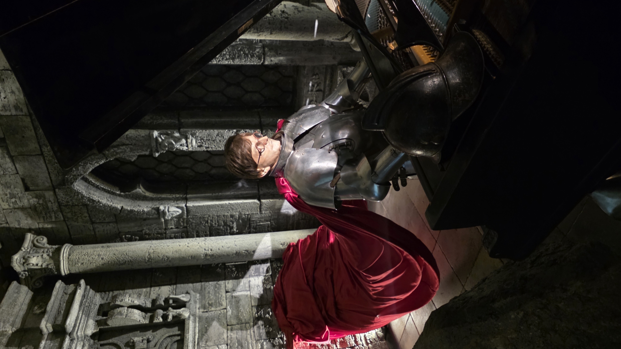 The image depicts a scene in what appears to be a medieval or historical setting. A wax figure of a man, seemingly dressed in silver knight armor, is positioned against a stone wall with gothic architectural elements. The figure wears glasses and has a red draped cloth extending from the armor, pooling onto the ground. The lighting is dim, with strong contrasts between light and shadow. The location suggests a museum or historical exhibit, potentially in Saint Petersburg, Russia, given the architectural style common in the city.
