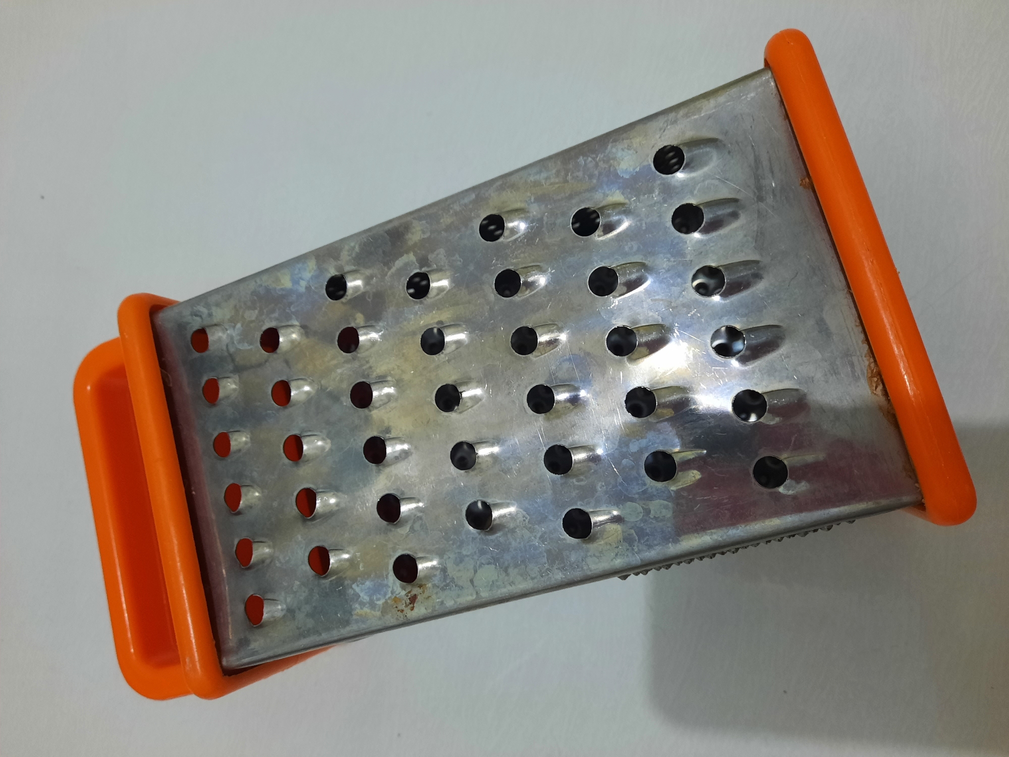 A close-up, high-angle, studio shot features an orange-handled, four-sided metal grater lying on a white, textured surface. The grater is positioned diagonally, with the top-left corner closest to the viewer. The metal surfaces show signs of wear and tear, with some discoloration and minor rust spots visible. Each side of the grater has different sized holes for grating, with some sides exhibiting finer grating teeth along the edges. The orange handles on the top and bottom are smooth and rounded. There are no people or other objects present in the scene. This image was captured in Lhokseumawe, Indonesia.